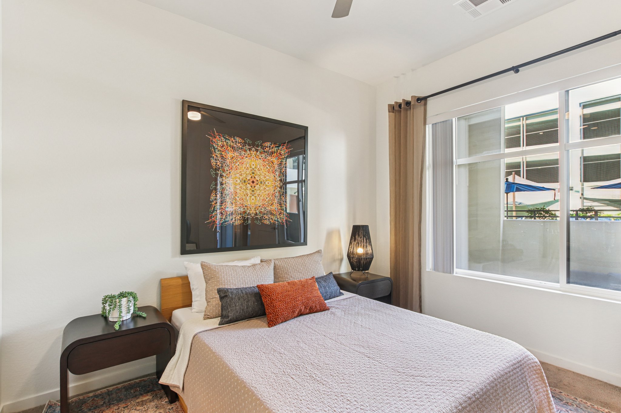 A well-decorated bedroom featuring a bed with a light-colored quilt, a bedside table, and a modern lamp. The wall behind the bed displays an abstract piece of art. Large windows allow natural light, with sheer curtains and a view of outdoor patio furniture. The space has a clean, contemporary aesthetic.