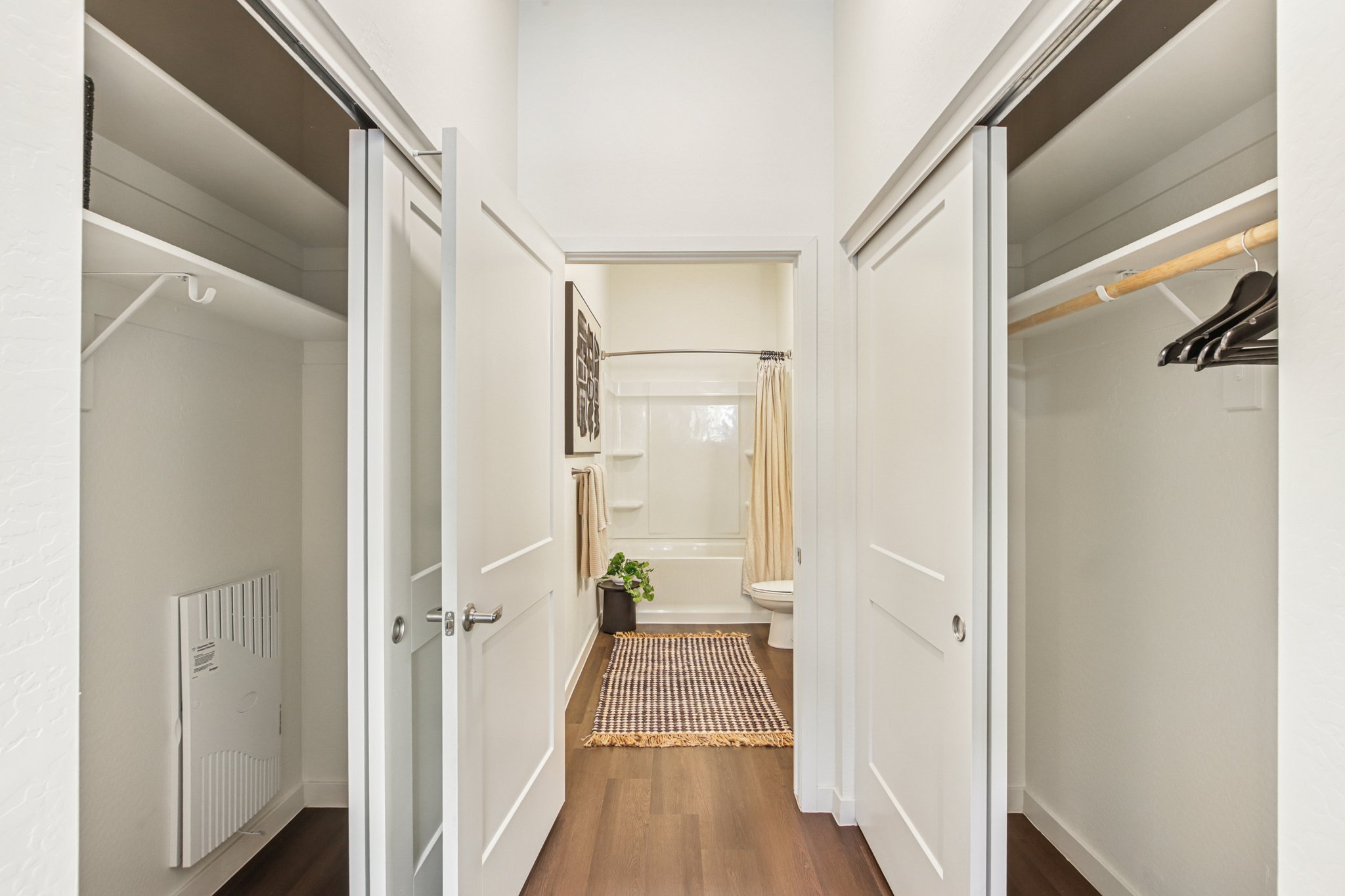 A view of a hallway with two open closet doors on either side and a bathroom at the end. The closets have white shelves and hangers, while the bathroom features a shower curtain, towels, and a small plant on the floor. The hallway has a wood floor and light-colored walls.