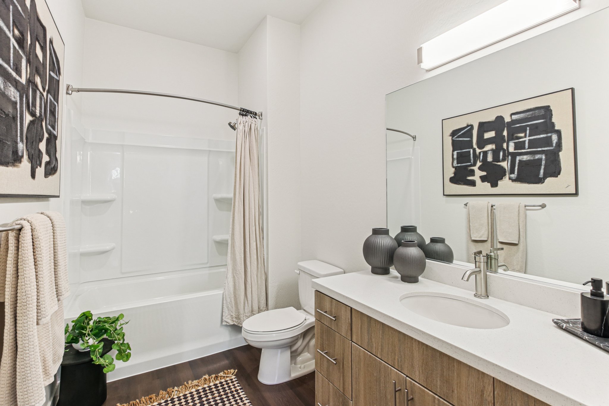 A modern bathroom featuring a white bathtub with a shower curtain, a round mirror above a sink with a wooden vanity, and decorative vases. Beige towels hang on a rack, and a green plant adds a touch of nature. The decor includes a framed artwork with abstract black designs on a light wall.