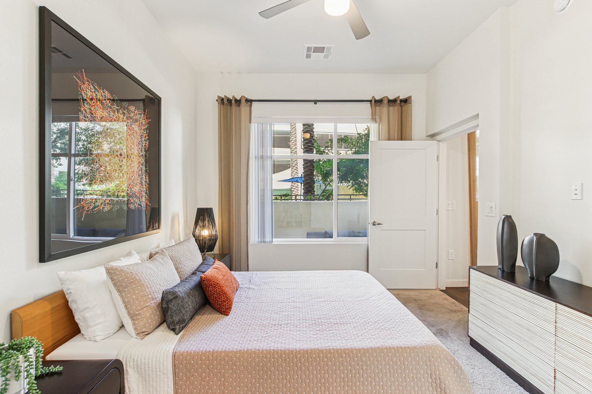 A modern bedroom featuring a queen-sized bed with decorative pillows, a sleek nightstand, and a stylish dresser. Large windows allow natural light to fill the room, showcasing a view of palm trees. The walls are painted in soft colors, complemented by contemporary artwork and warm lighting fixtures.