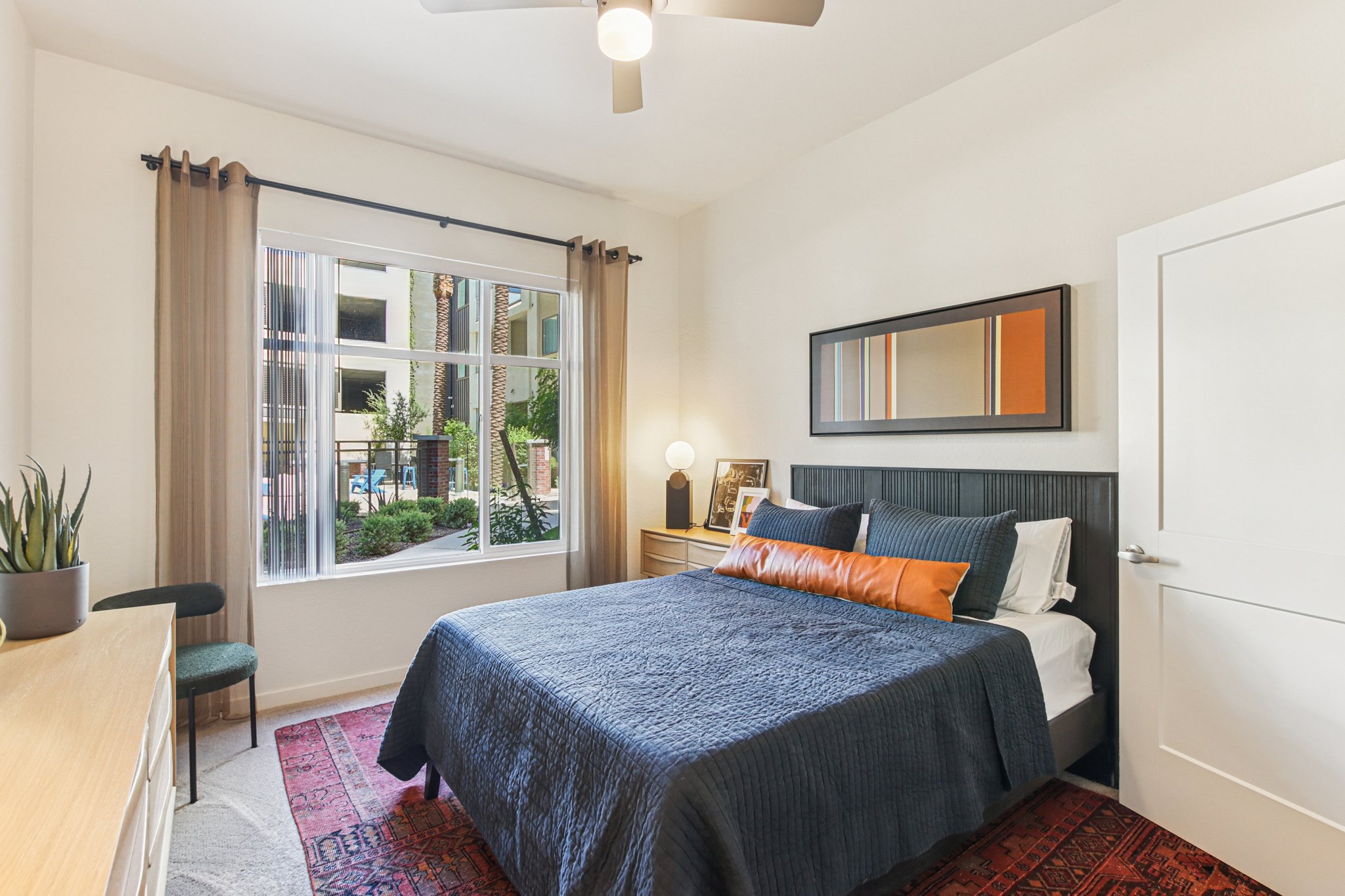 A cozy bedroom featuring a neatly made bed with dark blue bedding and an orange accent pillow. A nightstand with a lamp is beside the bed, and a large window lets in natural light, revealing greenery outside. A decorative rug and a plant add warmth to the space, creating a welcoming atmosphere.
