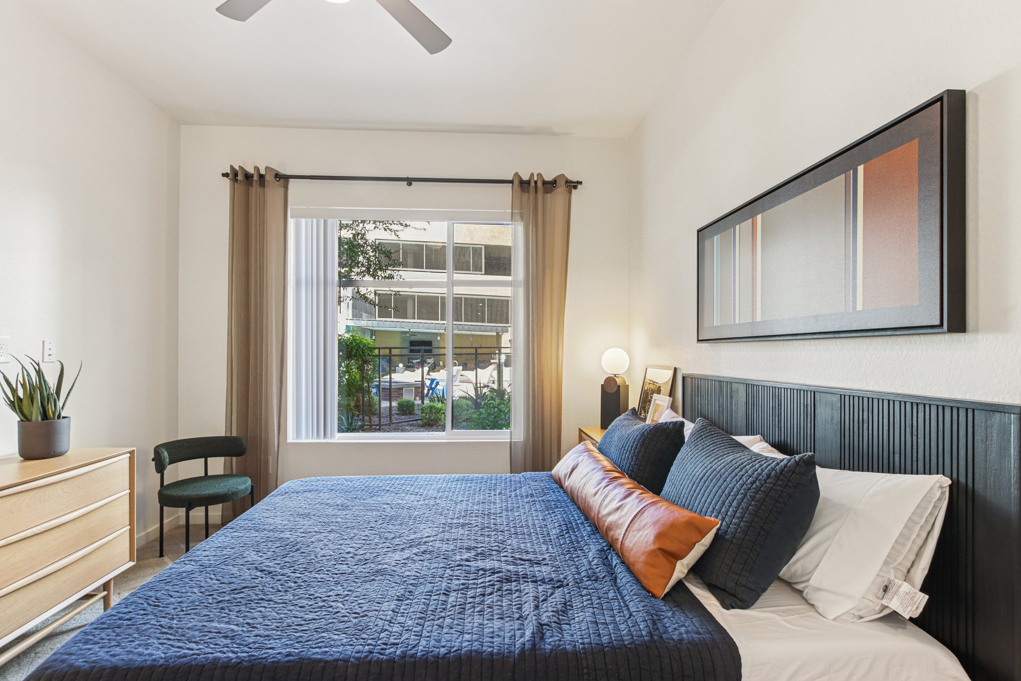 A modern bedroom featuring a neatly made bed with a dark blue quilt, decorative pillows, and a wooden headboard. A dresser is positioned beside the bed, and a green chair is visible. Large windows with sheer curtains allow natural light to enter, revealing a view of greenery outside.