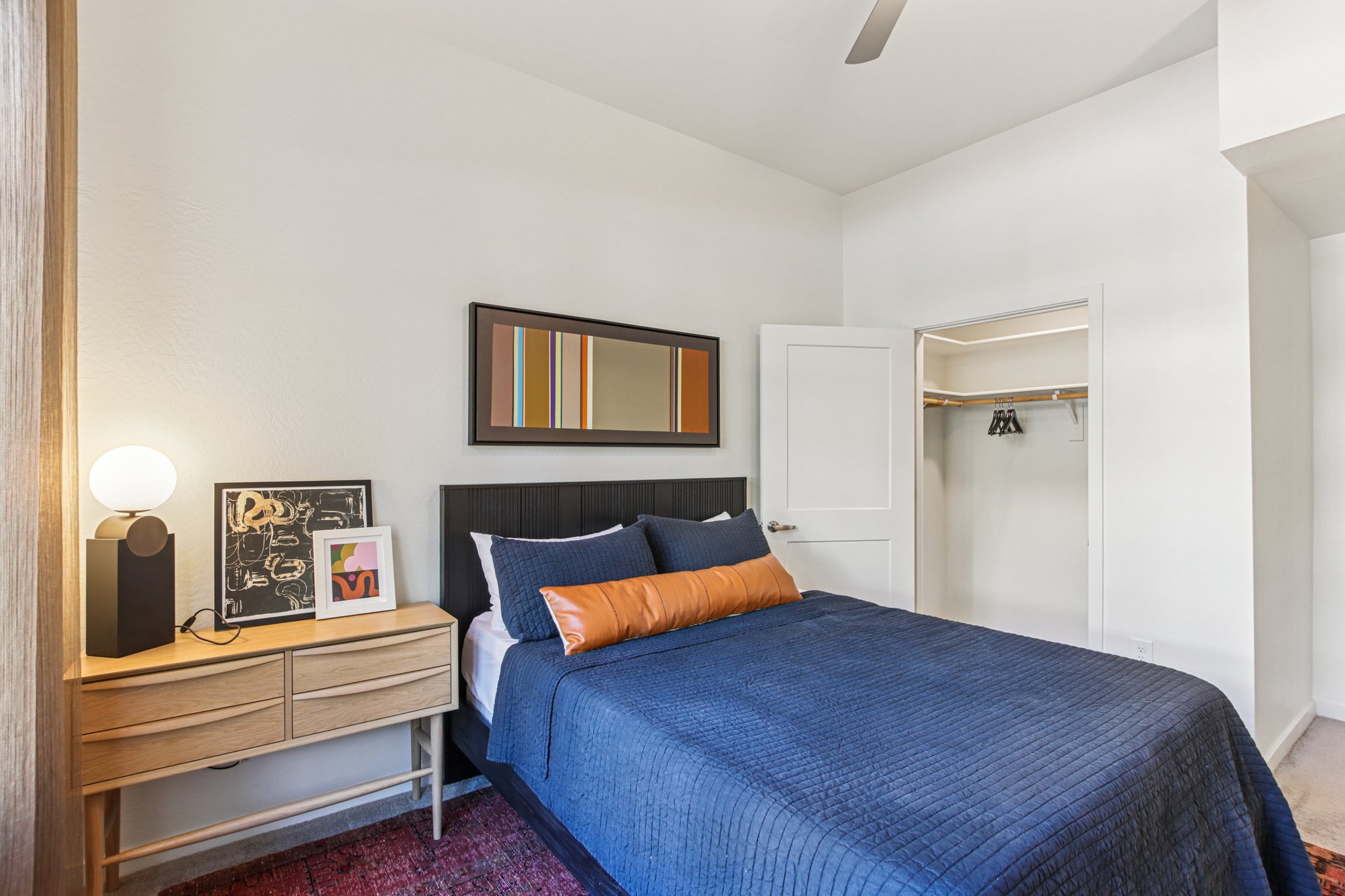 A well-decorated bedroom featuring a bed with a blue quilt, an orange pillow, a nightstand with a lamp and artwork, and a closet with open doors. The walls are painted white, and there is a modern mirror above the bed. A ceiling fan is visible, adding to the contemporary feel of the space.