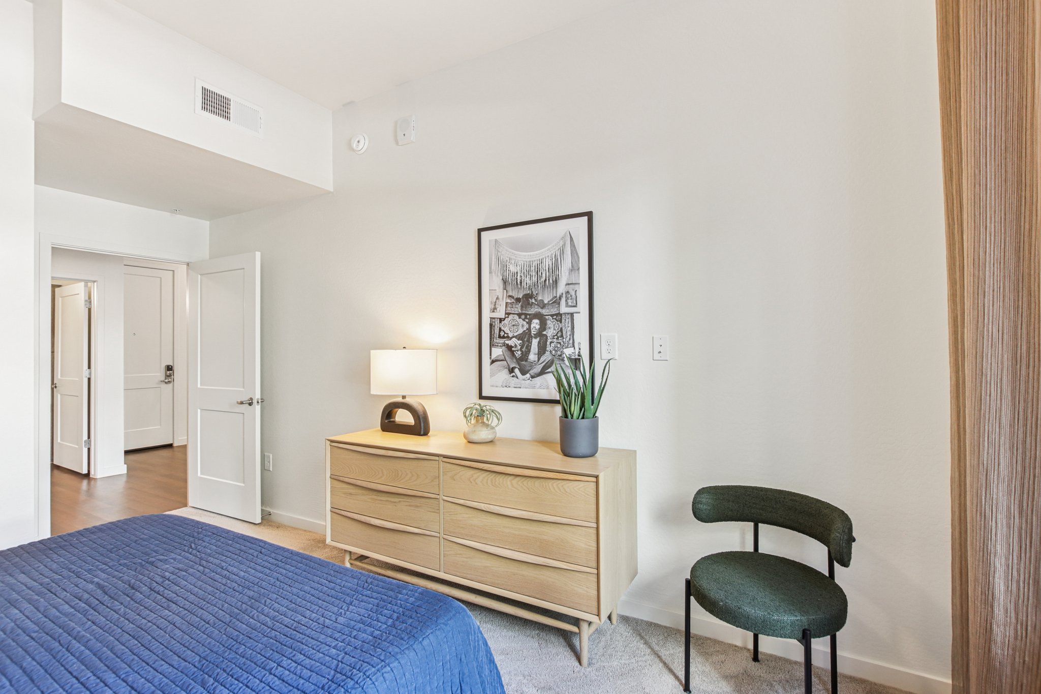 Cozy bedroom featuring a navy blue bedspread, a wooden dresser with a lamp and a potted plant, and a green chair. A framed artwork adorns the wall above the dresser, and a doorway leads to another room, highlighting a modern and inviting design. Natural light brightens the space.