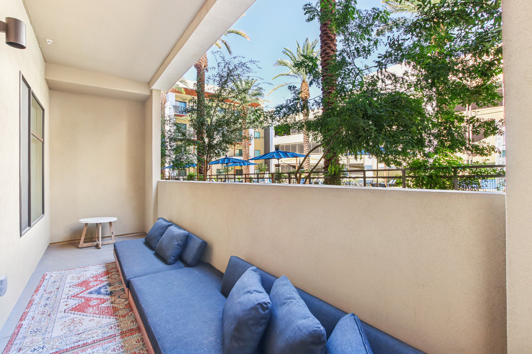 A modern patio area featuring a long, cushioned seating space with blue pillows, a small white table, and greenery surrounding the space. In the background, palm trees and blue umbrellas can be seen, indicating a pool or recreational area nearby. Bright and inviting atmosphere.