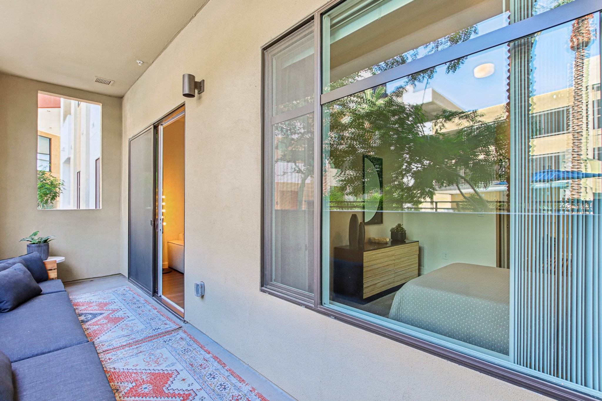 A cozy balcony with a gray sectional sofa and a patterned rug. Through large windows, a bedroom is visible, featuring a wooden dresser and a neatly made bed. Natural light filters in, highlighting the serene atmosphere of the outdoor space. Green plants are placed near the windows, enhancing the inviting ambiance.