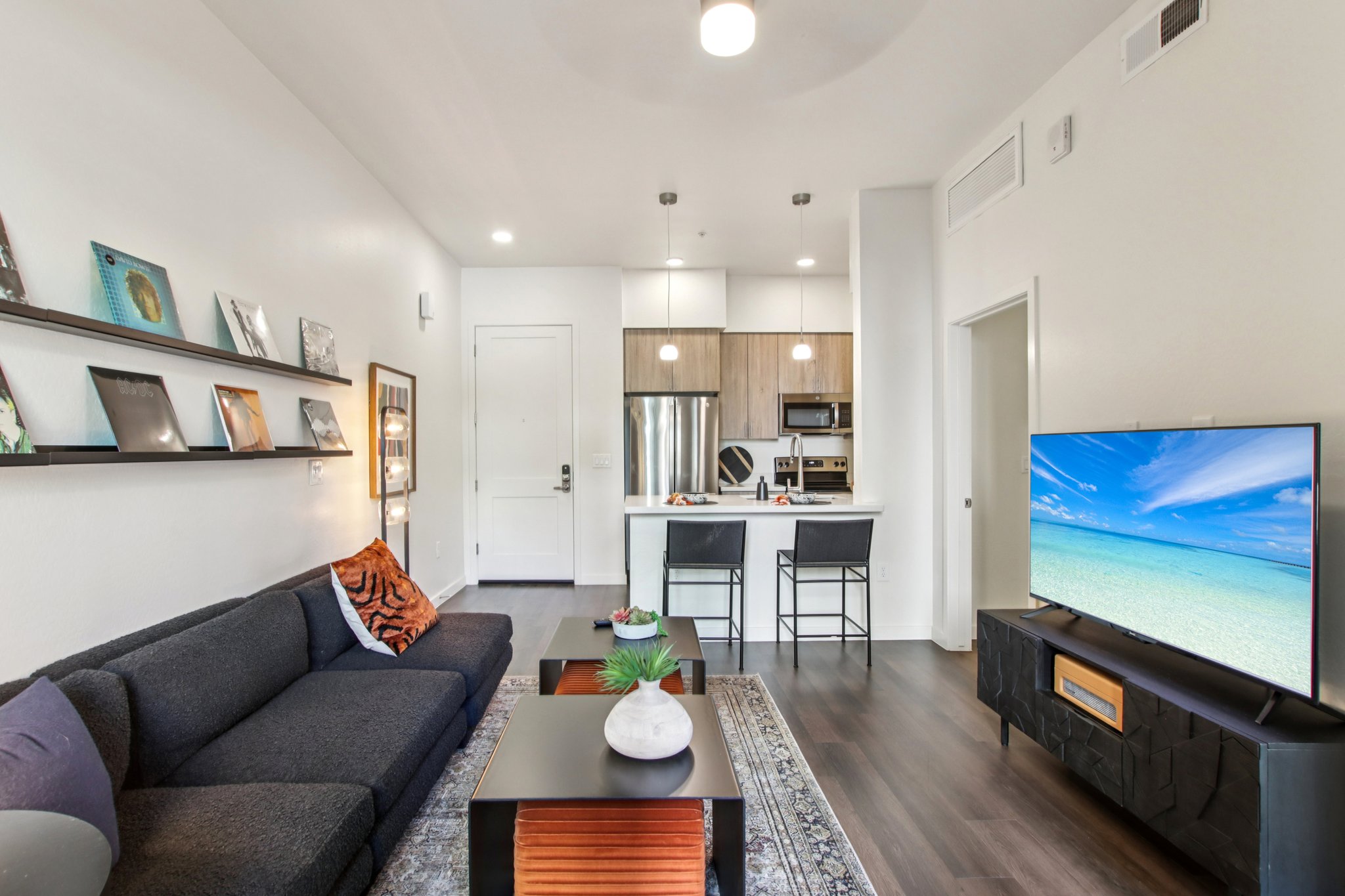 Modern living room featuring a dark sectional sofa with orange and black accent pillows, a coffee table with a small plant, and a television displaying a beach scene. The open layout includes a kitchen with wooden cabinetry and bar stools, and decorative wall art on shelves.