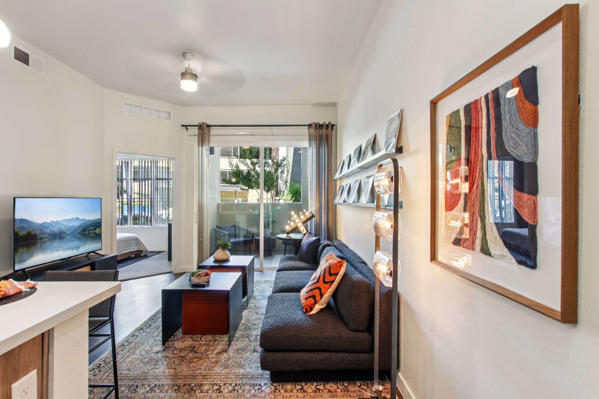 A modern living room featuring a gray sofa with an orange accent pillow, an abstract wall art piece, a coffee table, and a television. Large windows allow natural light, with a view of an outdoor area. Stylish lighting fixtures add warmth to the space, creating a cozy, inviting atmosphere.
