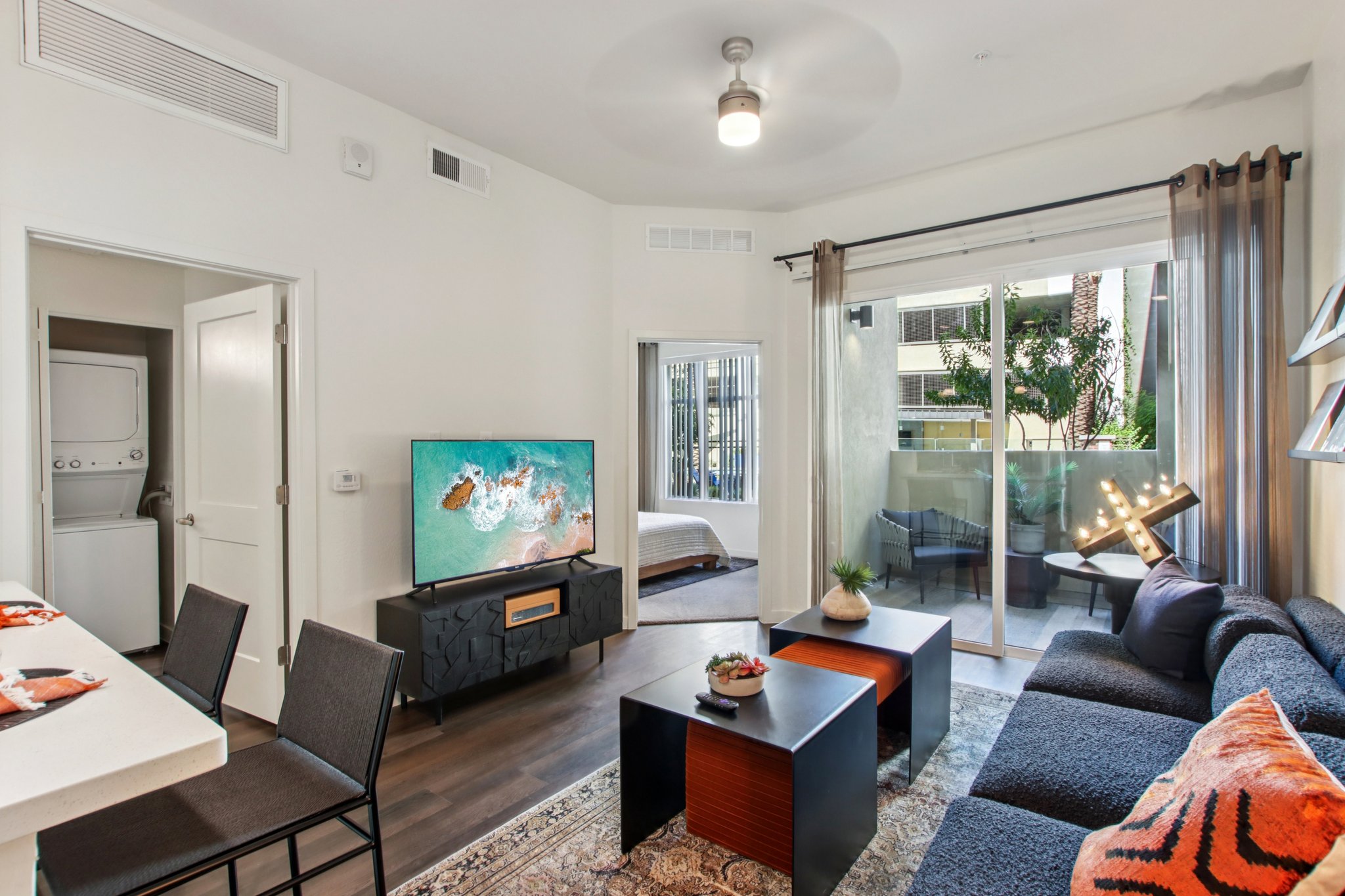 A modern living room featuring a dark sofa, glass coffee tables, and a large TV on a stylish cabinet. A dining area with four chairs is visible, along with a door leading to a bedroom and a sliding glass door opening to a balcony with greenery. Bright, contemporary decor enhances the space.