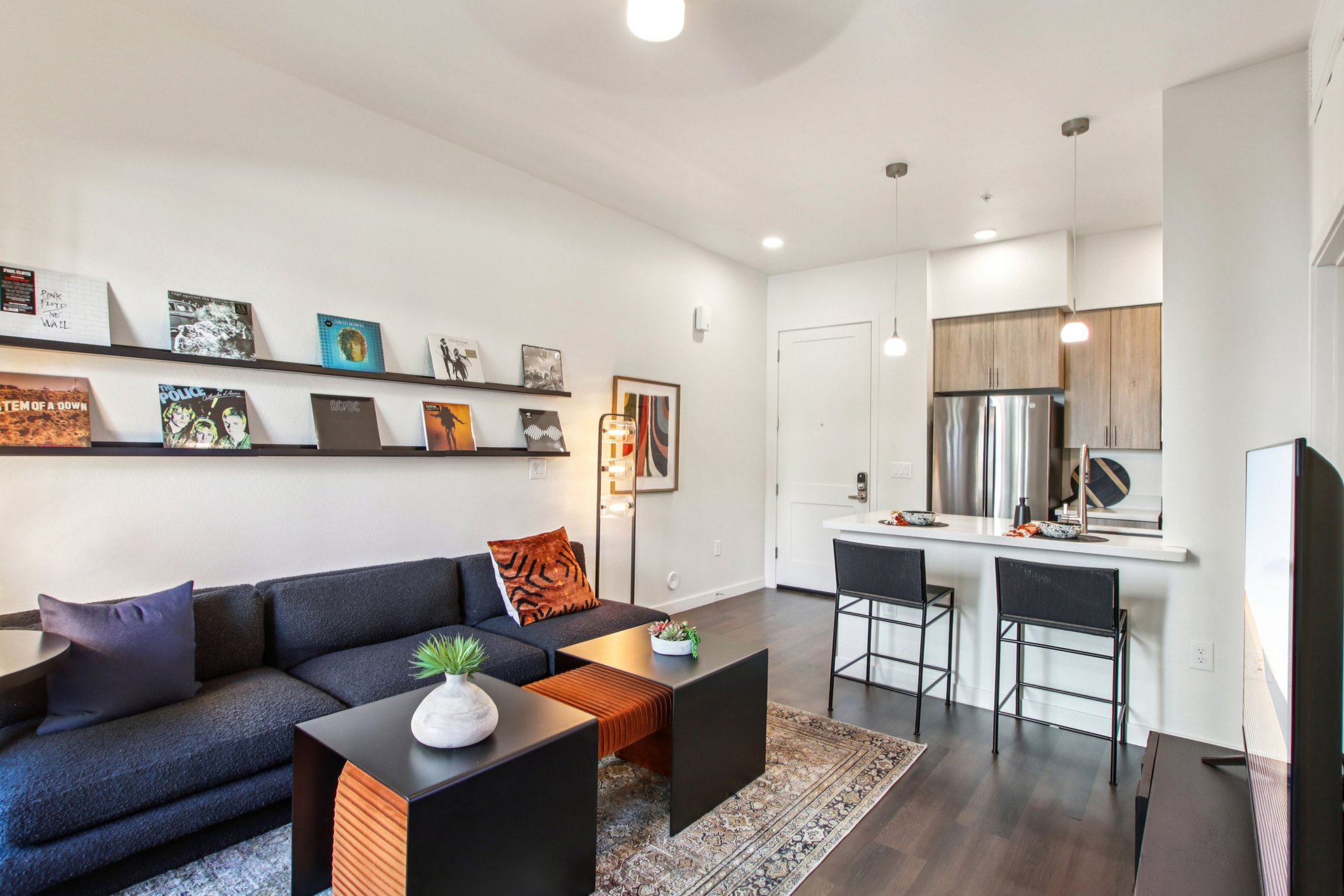 A modern living room with a navy blue sofa and two black coffee tables. Wall shelves display vinyl records and artwork. A small dining area with two black chairs and a kitchen in the background featuring stainless steel appliances and wood cabinetry. Natural light illuminates the space.