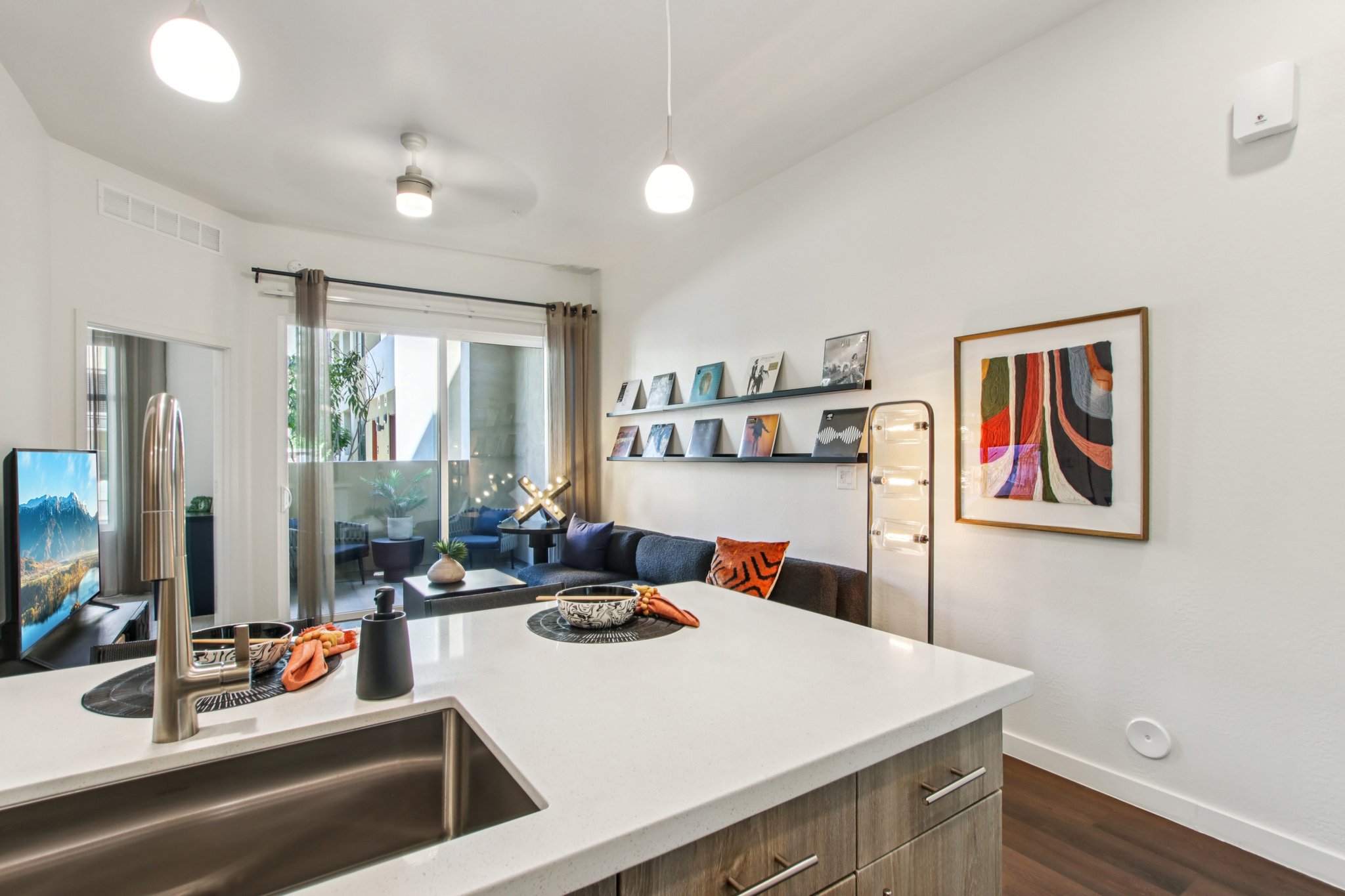 A modern apartment interior featuring a kitchen with a large island and sink, a cozy living area with a sectional couch, decorative wall art, and a television. There are bright lights and plants visible through large windows, creating a warm and inviting atmosphere.