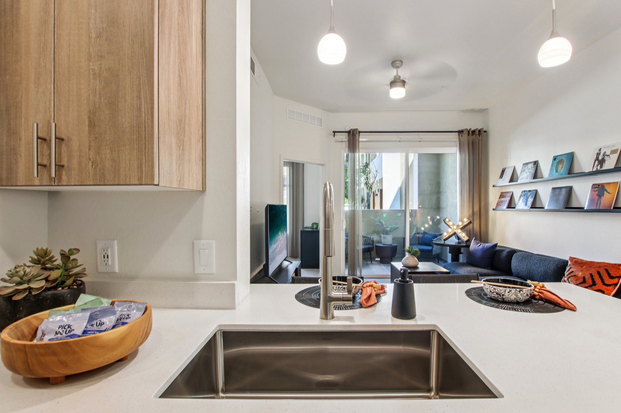 Modern kitchen with a sleek countertop and stainless steel sink. A wooden bowl and decorative dishes are placed on the counter. In the background, a cozy living area features a couch, a coffee table, and framed artwork on the wall, with natural light coming in from a glass door leading to an outdoor space.