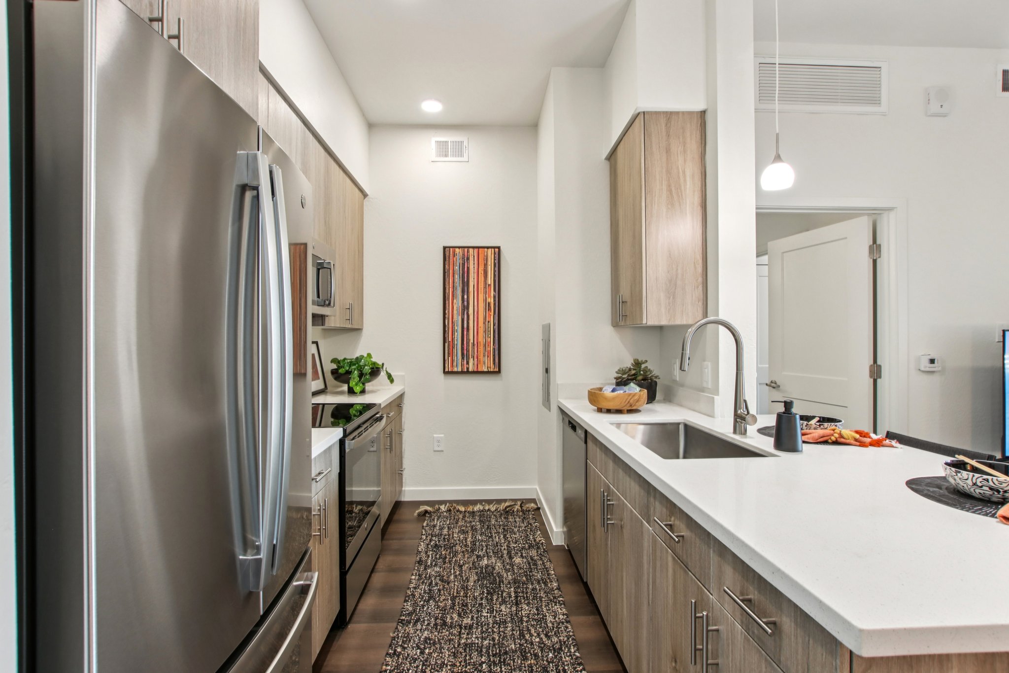 Modern kitchen featuring stainless steel appliances, light wood cabinets, and a white countertop. A small decorative rug runs along the floor, and a framed artwork is mounted on the wall. The space is bright and minimalistic, with an open layout leading to another room.