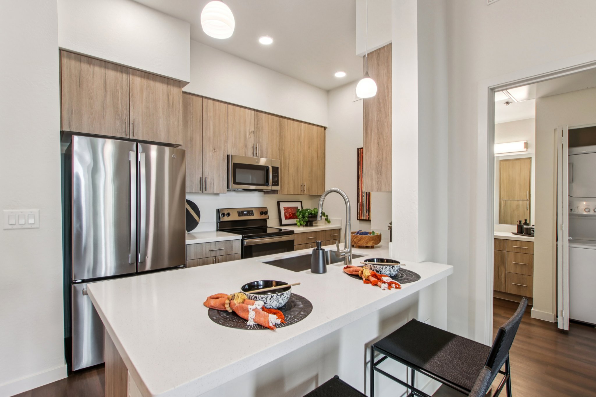 Modern kitchen featuring stainless steel appliances, including a refrigerator and microwave. Light wooden cabinets complement a white countertop. Two black bar stools with woven baskets sit at the island, which is set with two decorative bowls. A doorway leads to a laundry area, enhancing convenience.