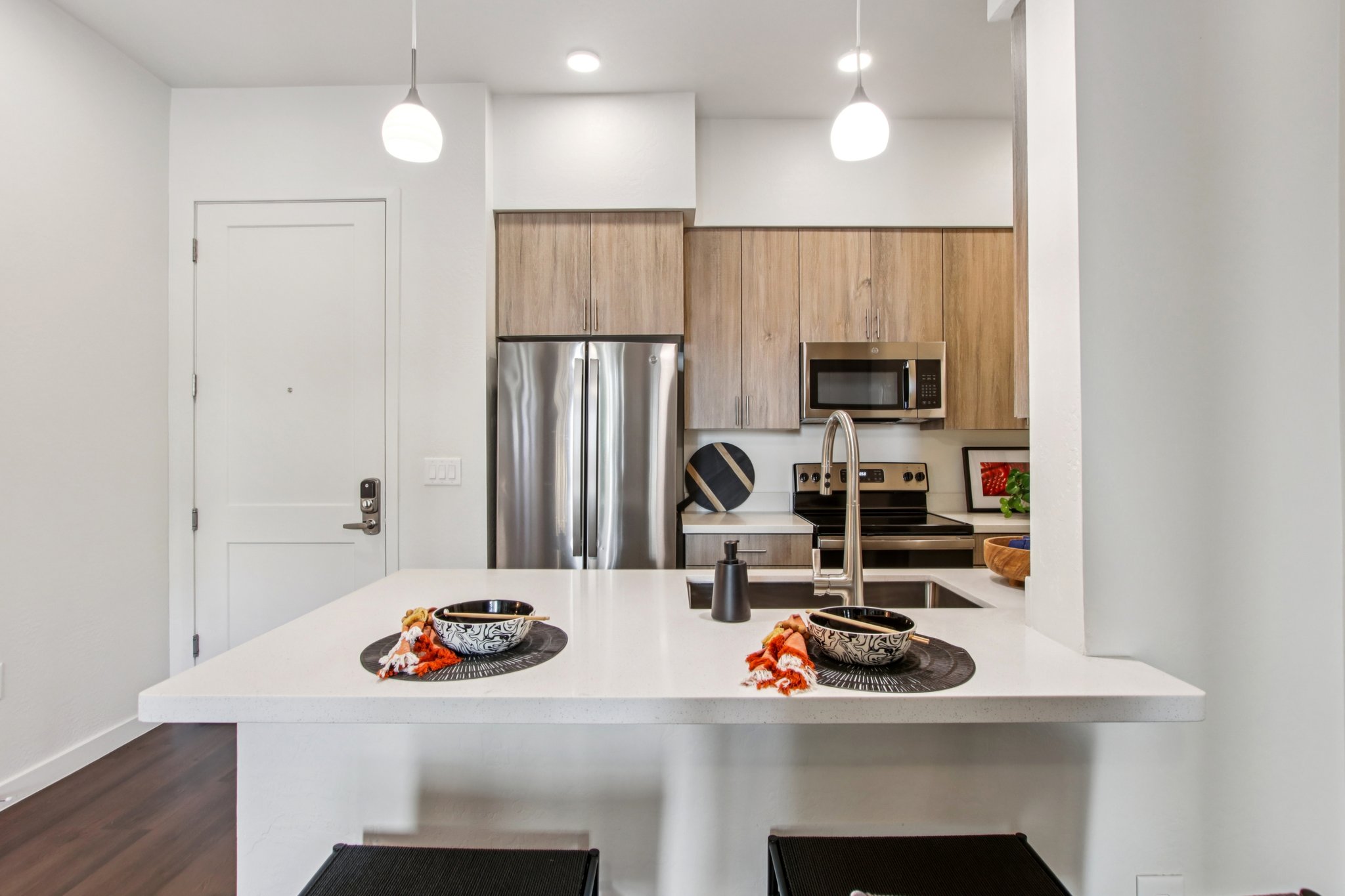 Modern kitchen featuring a white countertop with two black bowls, stainless steel appliances including a fridge and microwave, light wooden cabinetry, and pendant lighting. There are decorative elements like a small plant and a fruit bowl on the counter. The kitchen has a clean and contemporary design.
