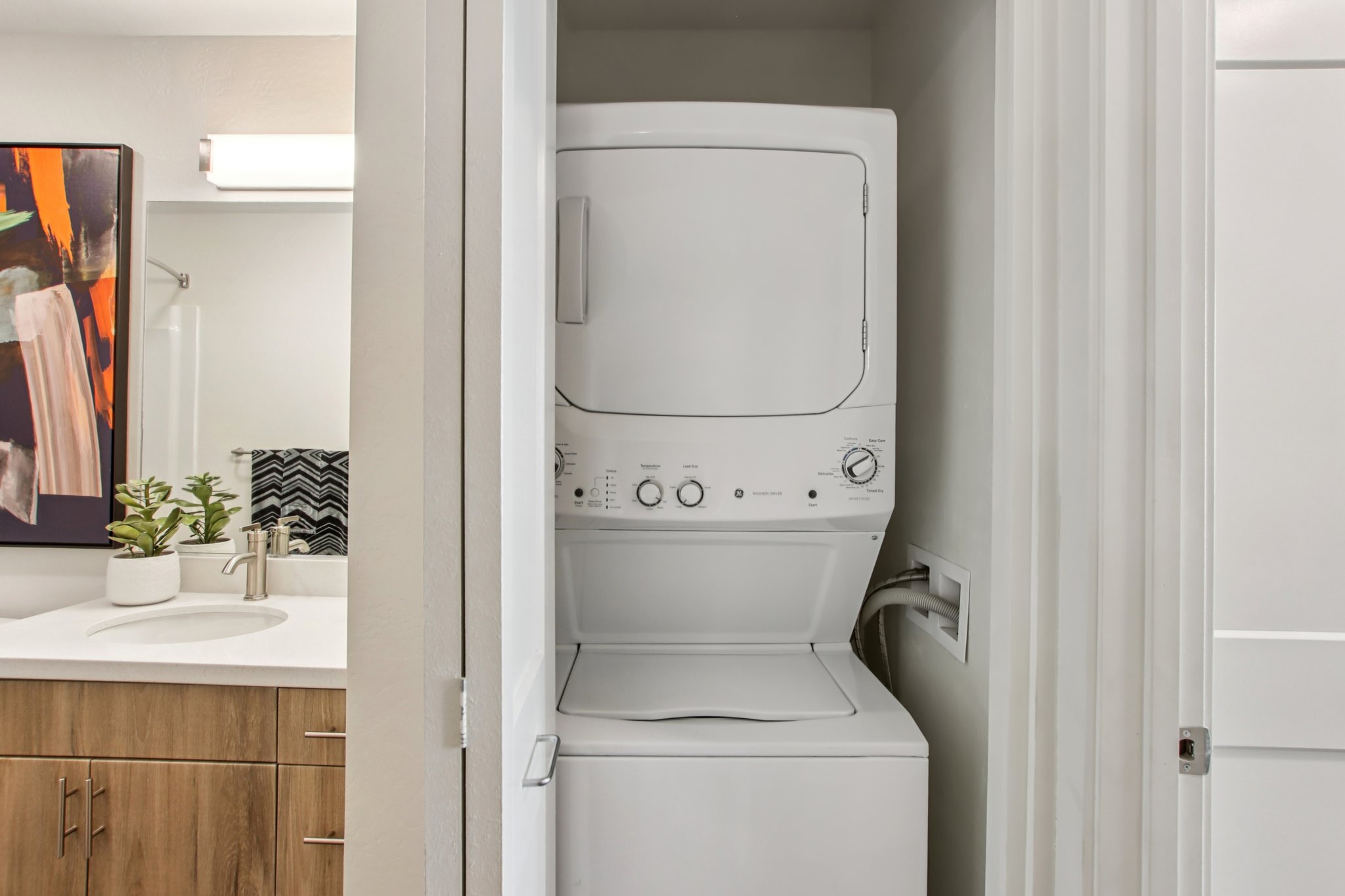 A compact laundry area featuring a stacked washer and dryer unit. The background includes a bathroom sink with a dark cabinetry and a decorative plant. A door is partially open, revealing the laundry appliances in a modern, clean setting.