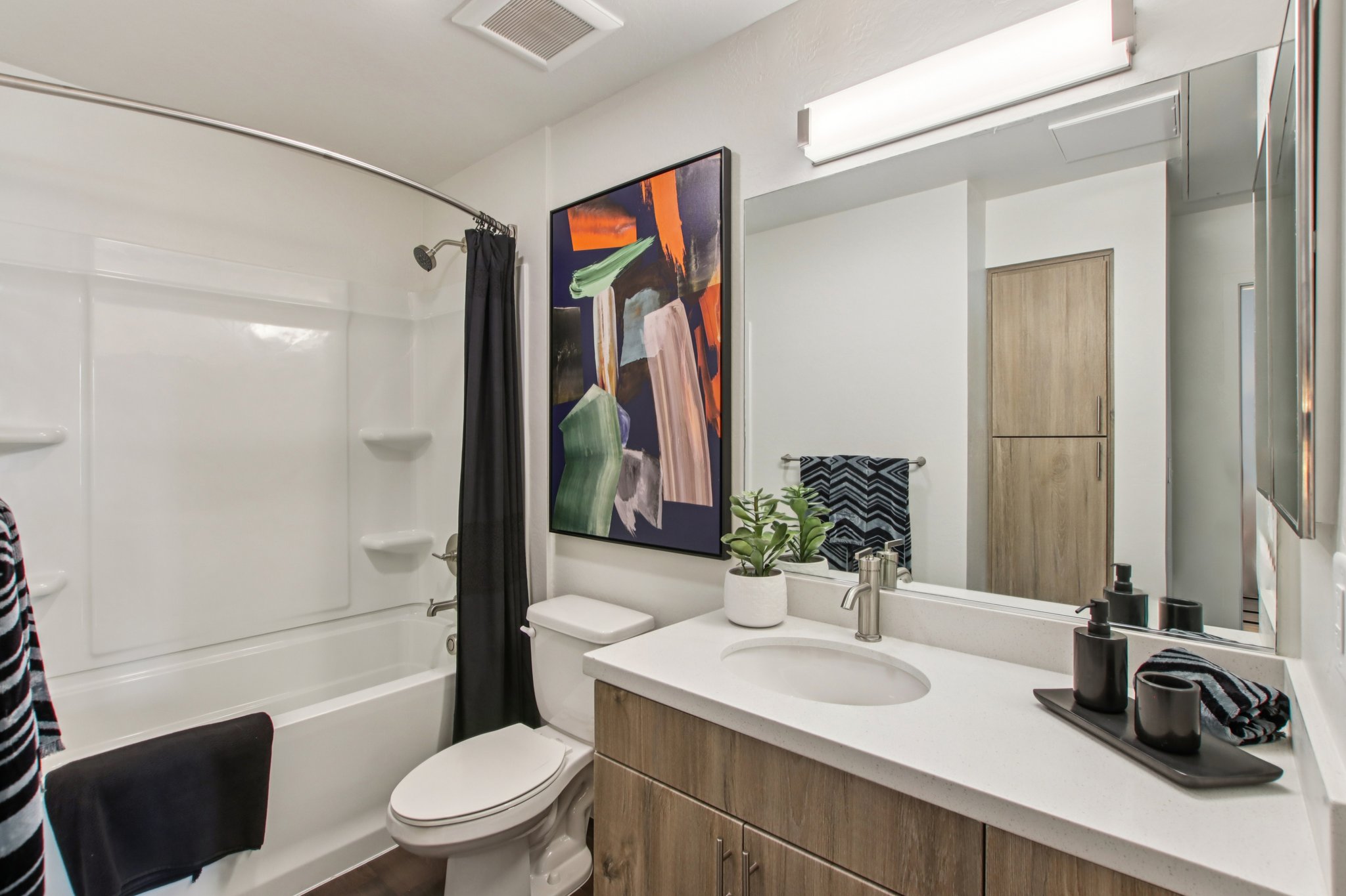 A modern bathroom featuring a white bathtub with a shower curtain, a white toilet, and a sleek wooden vanity. The vanity has a sink and organized toiletries. A colorful abstract painting hangs on the wall, and a small plant adds a touch of greenery. Natural light brightens the space.