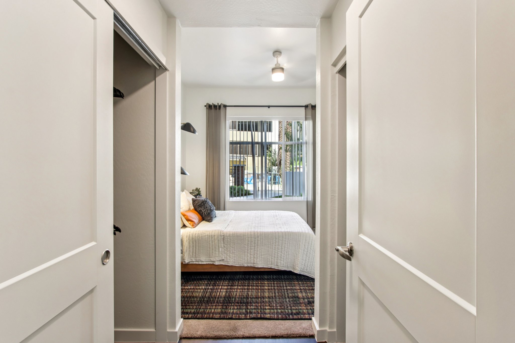 A well-lit, modern bedroom seen through an open doorway. The room features a neatly made bed with white bedding and an orange accent pillow. Natural light streams in through a window, which is dressed with light curtains. The hallway has white walls and a decorative rug on the floor.
