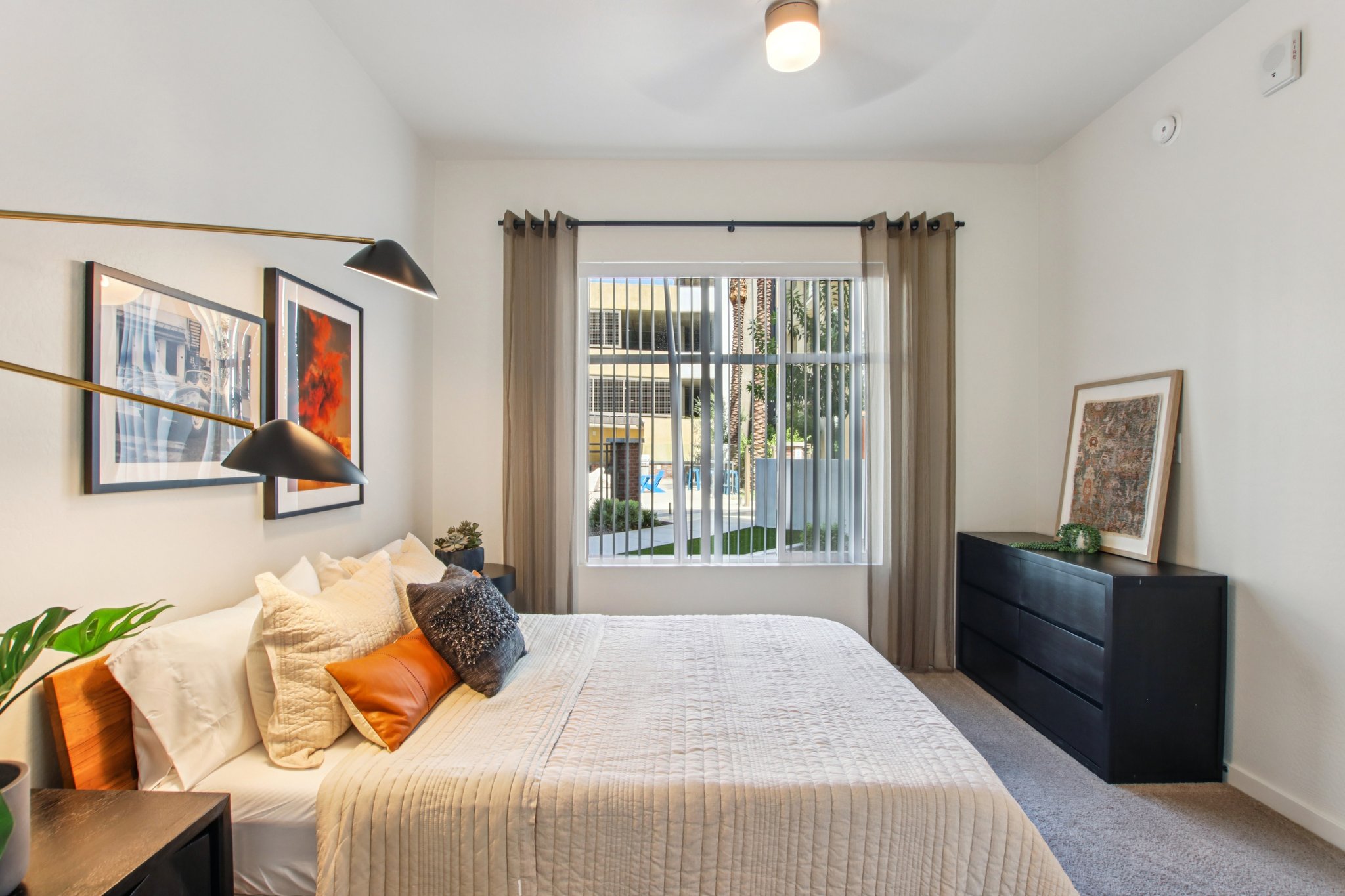 A modern bedroom featuring a queen-sized bed with decorative pillows, a nightstand with a plant and lamp, and a dark dresser. The room has large windows with sheer curtains, allowing natural light to enter, and artwork hanging on the walls, creating a cozy and inviting atmosphere.
