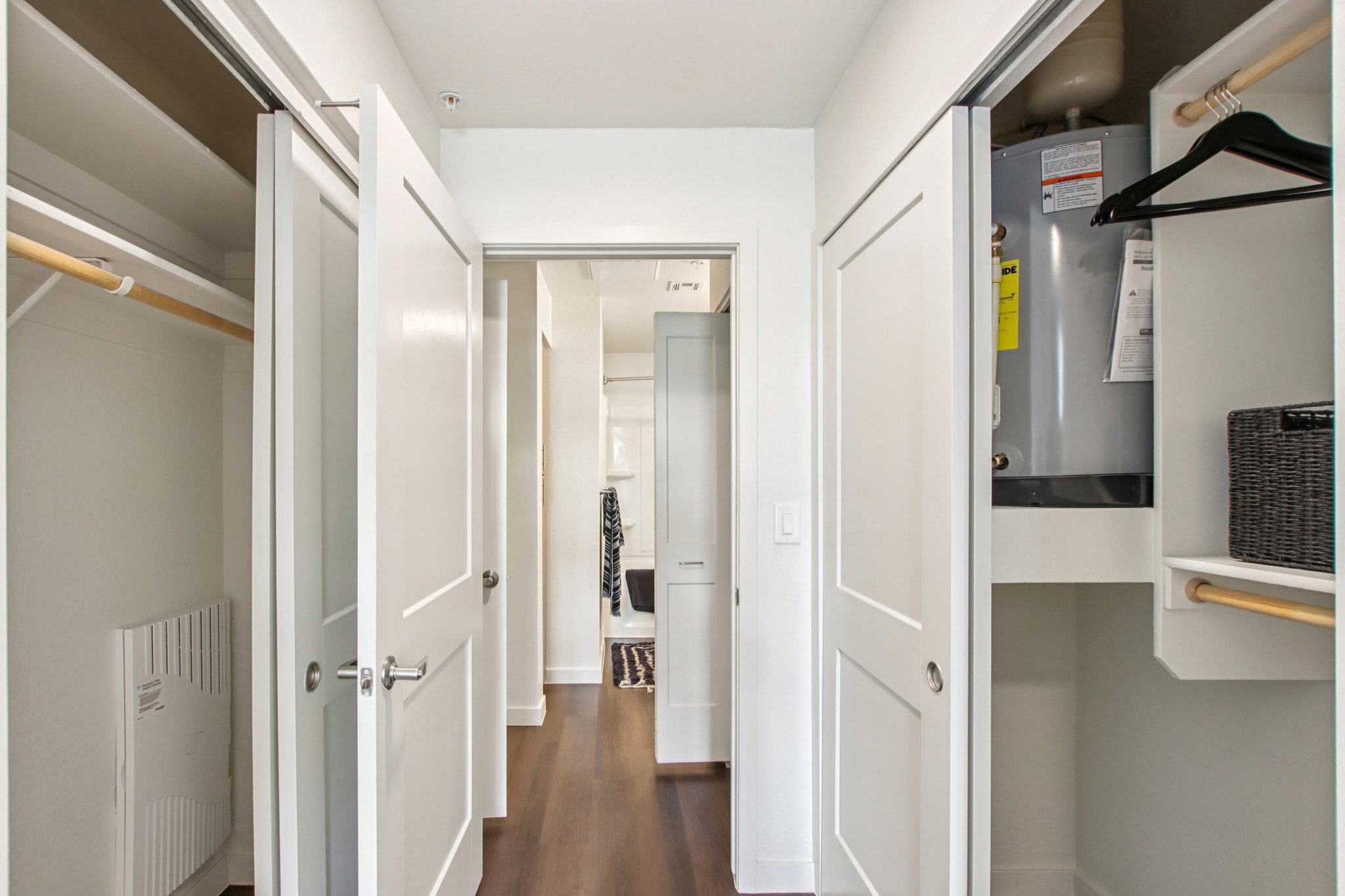 Interior view of a modern closet area featuring two open doors. One door leads to a small storage space with a hot water heater, while the other provides access to a hallway. The space is brightly lit with white walls and has wooden flooring. A black basket is visible on a shelf in the closet.