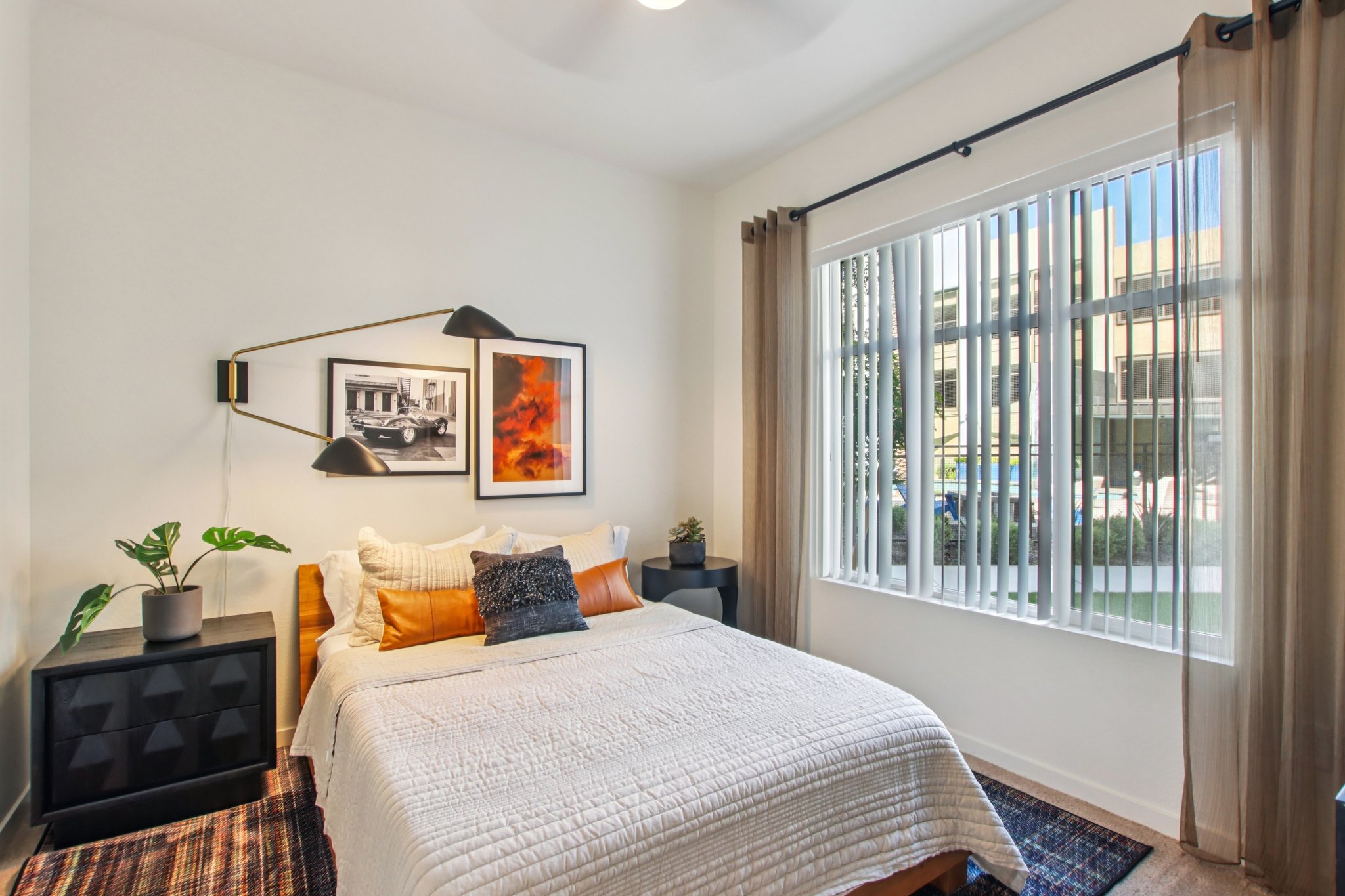 A cozy bedroom with a queen-sized bed dressed in white linens and decorative pillows. The room features a stylish wall lamp, a small side table, and a potted plant. Natural light filters through large windows dressed with sheer curtains, and framed artwork decorates the walls. The floor is covered with a patterned rug.