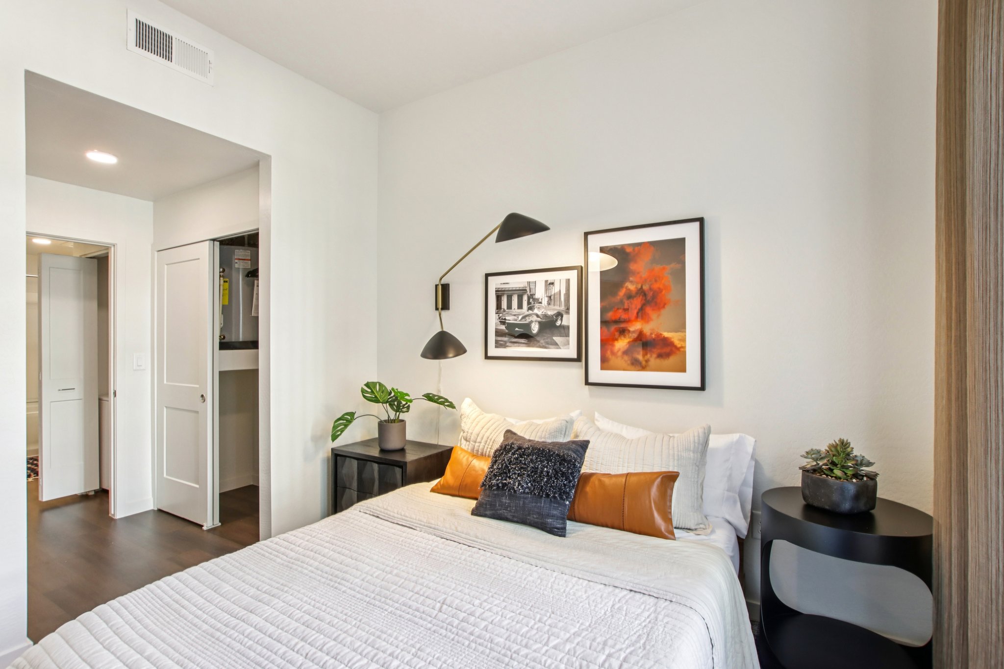 A cozy bedroom featuring a queen-sized bed with white bedding and decorative pillows. A modern lamp is mounted on the wall above the bed, and two framed artworks hang above it. A small black nightstand holds a plant and a decorative item. A closet is visible in the background, with light-colored walls and dark flooring.