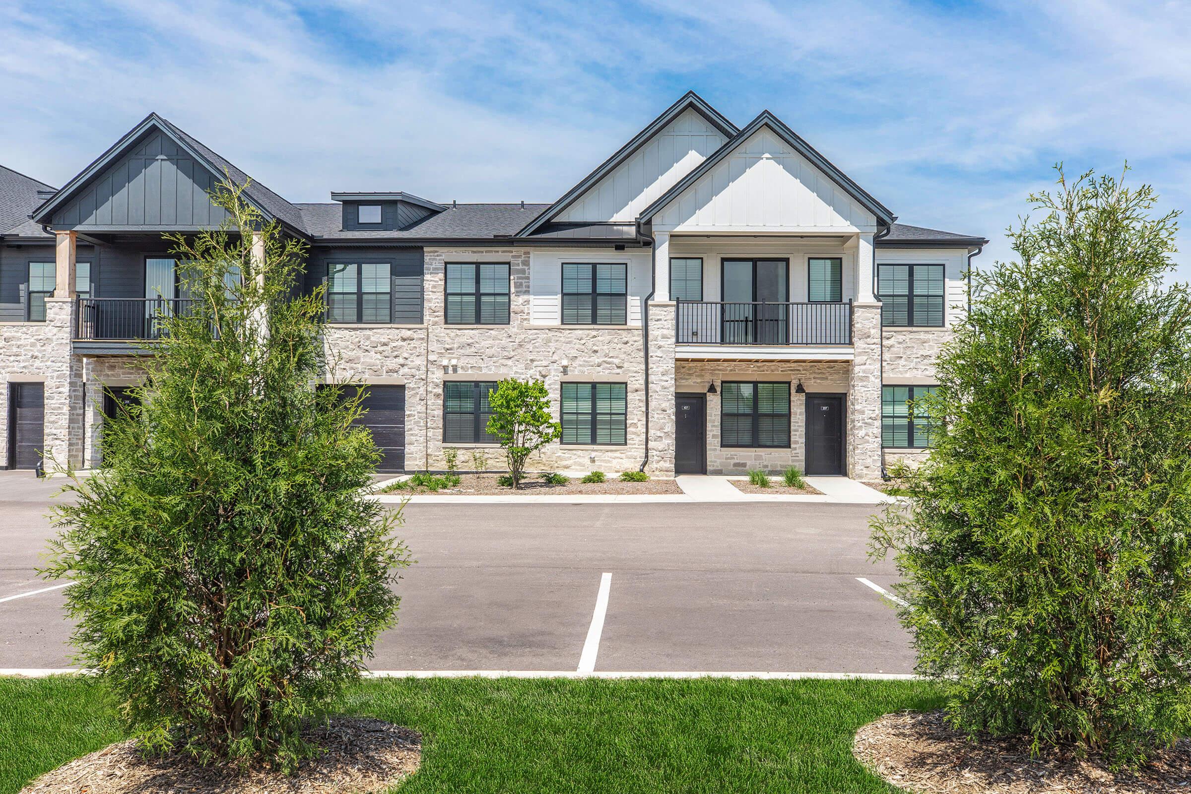A modern residential building featuring a combination of stone and siding, with multiple units visible. The facade has balconies, large windows, and a well-maintained landscape in the foreground, including trees and a parking area. The sky is bright and clear, suggesting a sunny day.