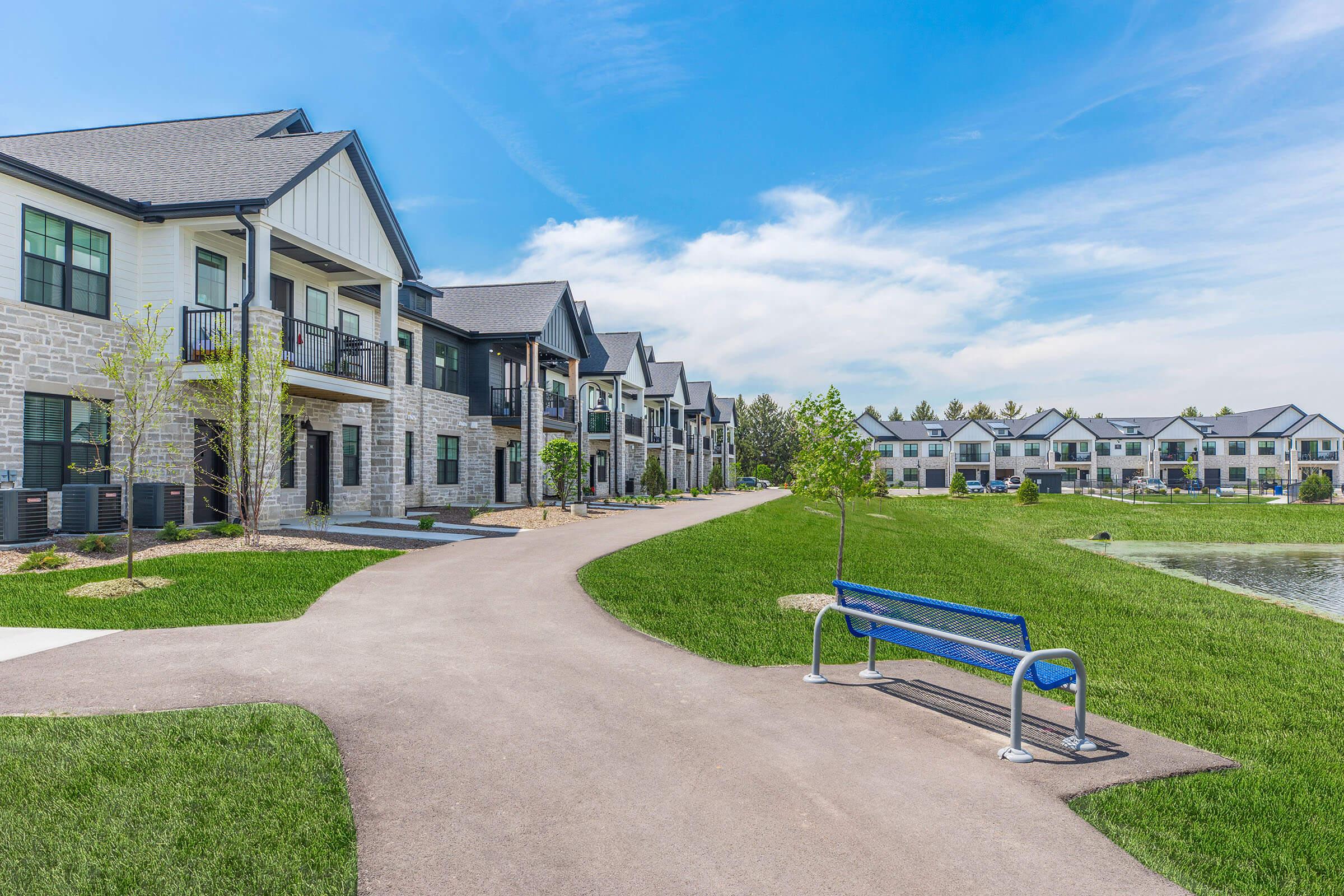 A scenic view of a modern residential complex featuring two-story buildings with a mix of stone and siding exteriors. A paved walkway winds through well-maintained green lawns, with a blue park bench placed nearby and a serene pond visible in the background under a clear blue sky.