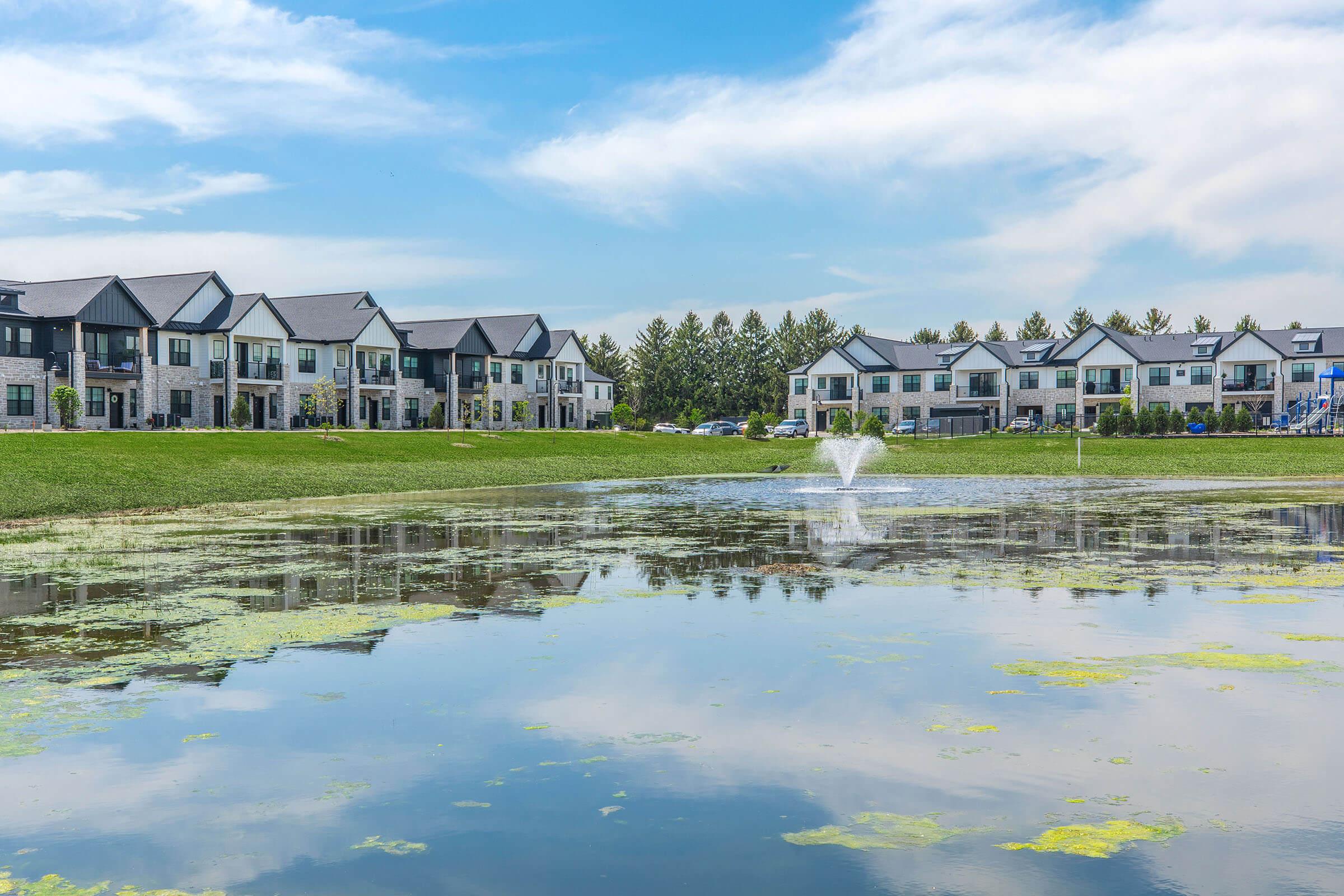 A serene landscape featuring a pond in the foreground with a fountain, surrounded by greenery. In the background, there are modern multi-story apartment buildings under a clear blue sky, reflecting in the water. The area appears well-maintained with lush grass and trees lining the property.