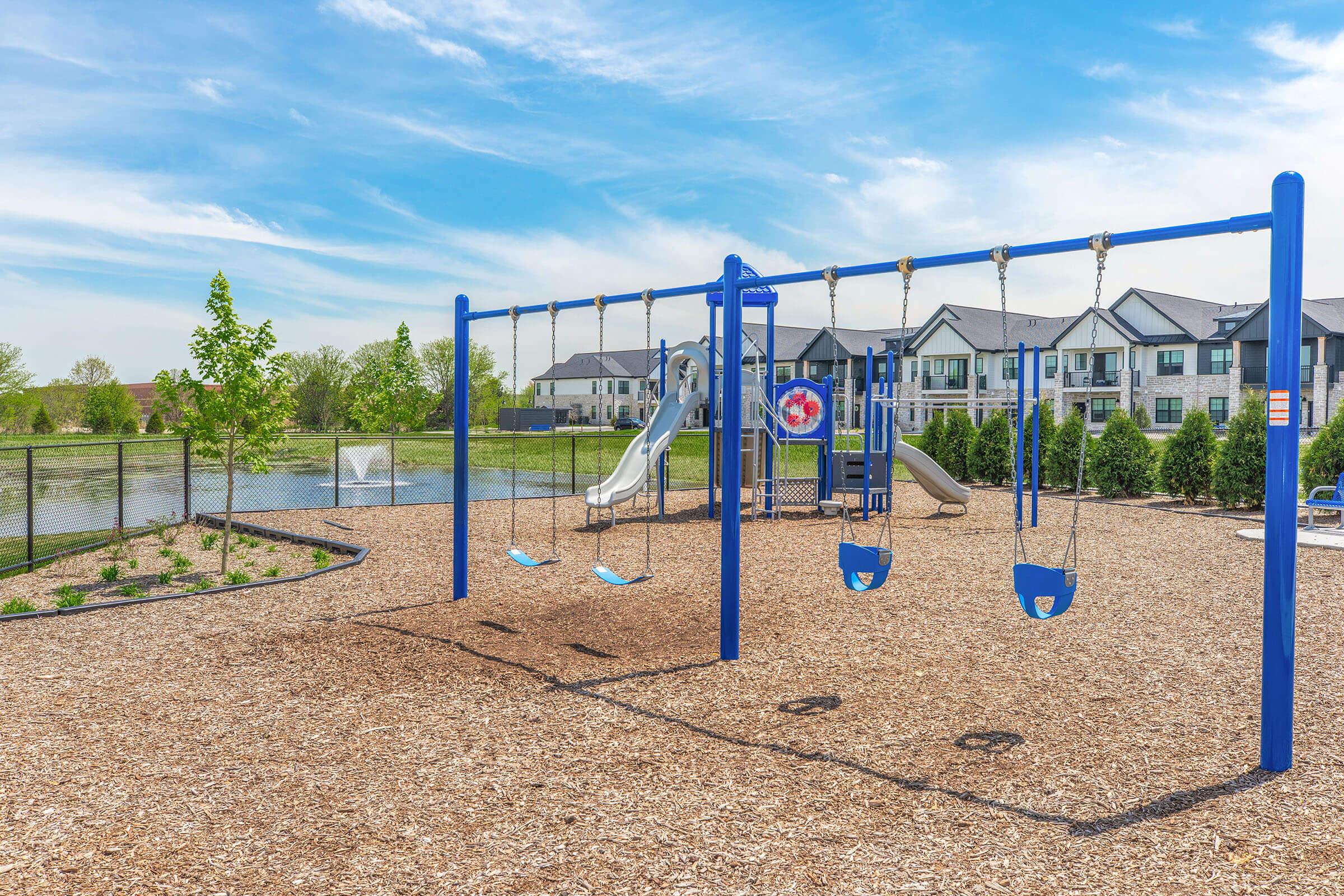 A playground featuring two blue swings and a slide, surrounded by soft wood chip flooring. In the background, there are several modern apartment buildings and a pond, with green trees and blue skies creating a pleasant outdoor setting.