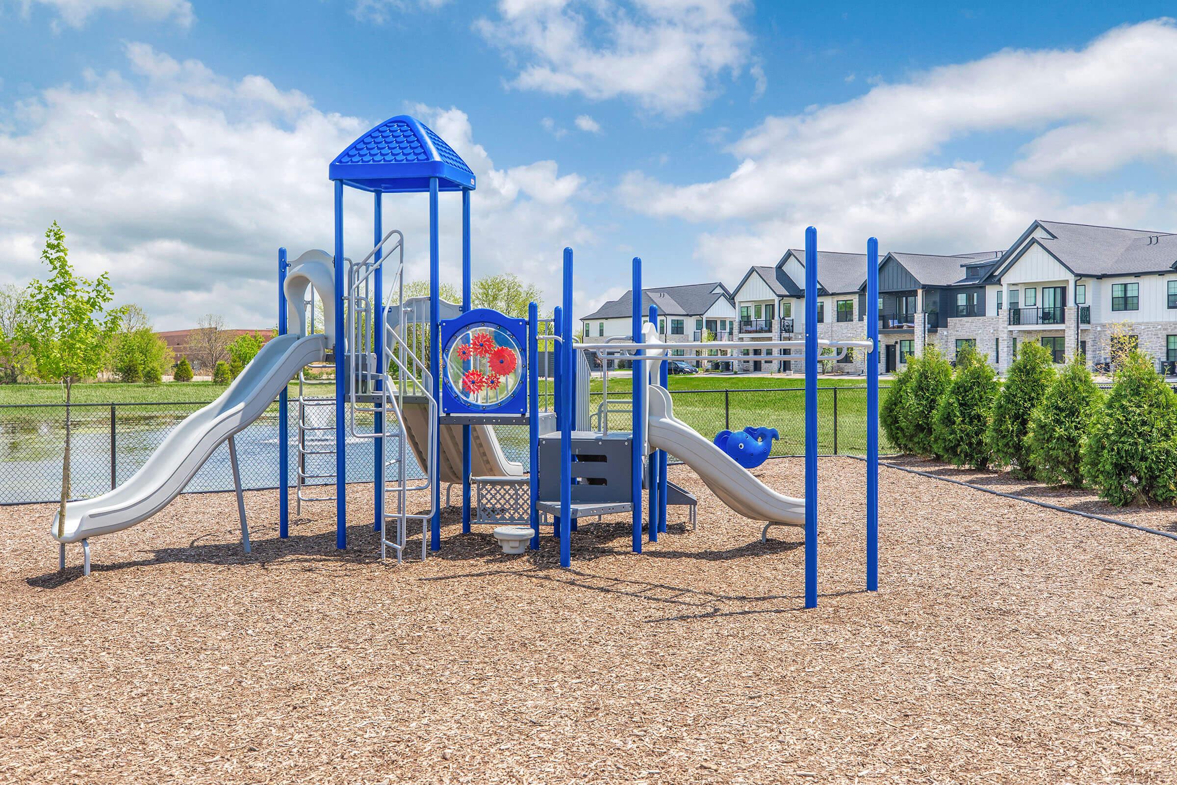 Bright playground equipment featuring two slides, a climbing structure, and a play panel, set in a sunny outdoor area with wood mulch. In the background, there are multi-story residential buildings and a lush green landscape under a partly cloudy sky.