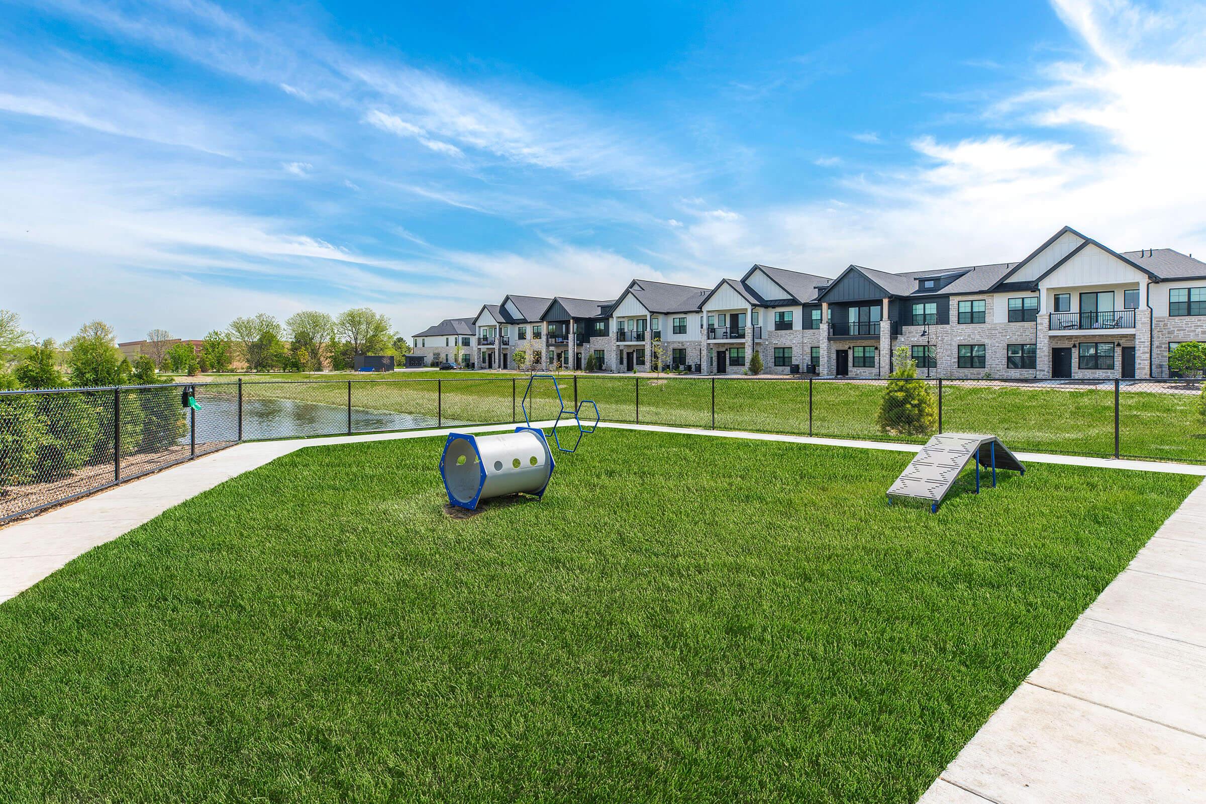 A spacious park area featuring bright green grass, dog agility equipment including a tunnel and a ramp, and a fenced section. In the background, there are modern residential buildings and a calm pond under a clear blue sky.