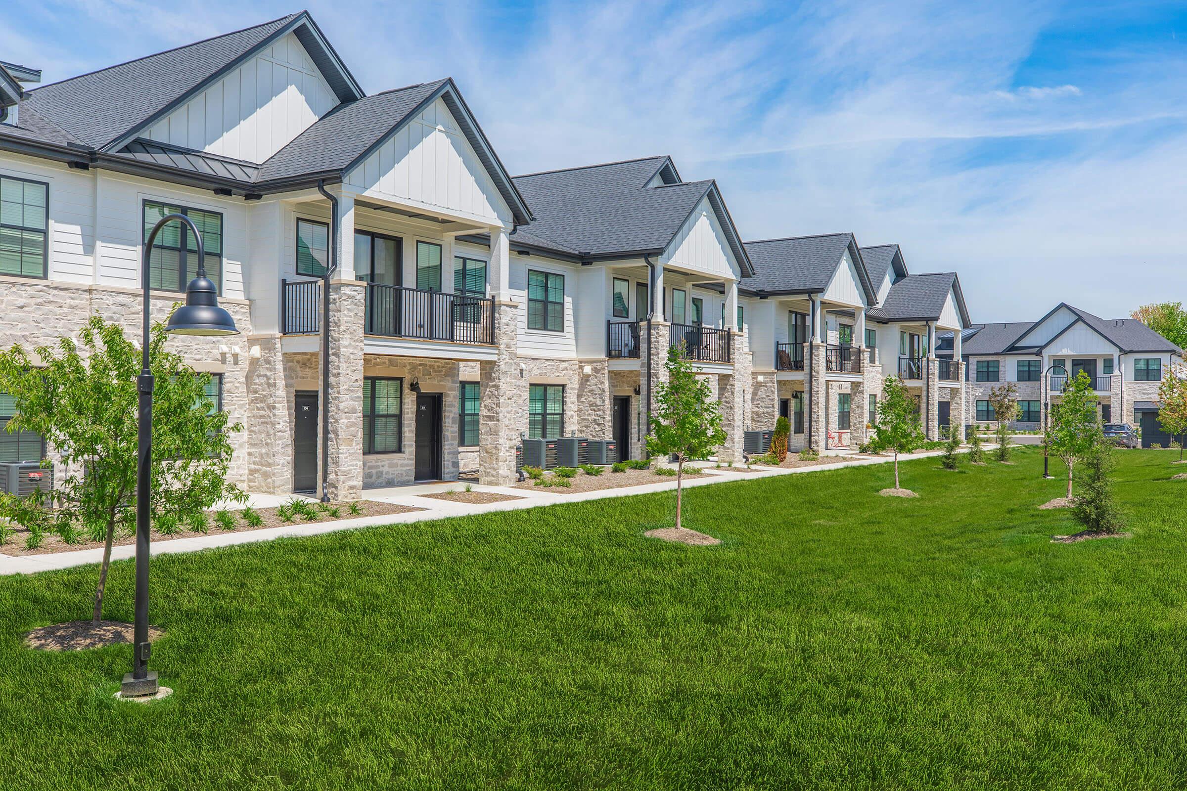 A row of modern apartment buildings with stone and siding exteriors, featuring balconies and windows. The well-maintained grassy area in front has small trees and a pathway, creating a pleasant outdoor environment. The sky is clear with a few clouds.