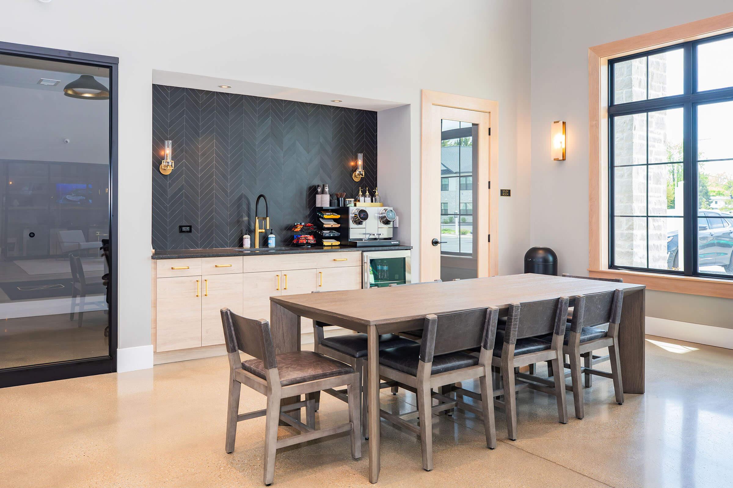 A modern break room featuring a wooden dining table with six chairs, a sleek coffee station with various appliances, a textured wall backdrop, and large windows allowing natural light. The space has a contemporary design with neutral colors and stylish lighting, creating a welcoming atmosphere.
