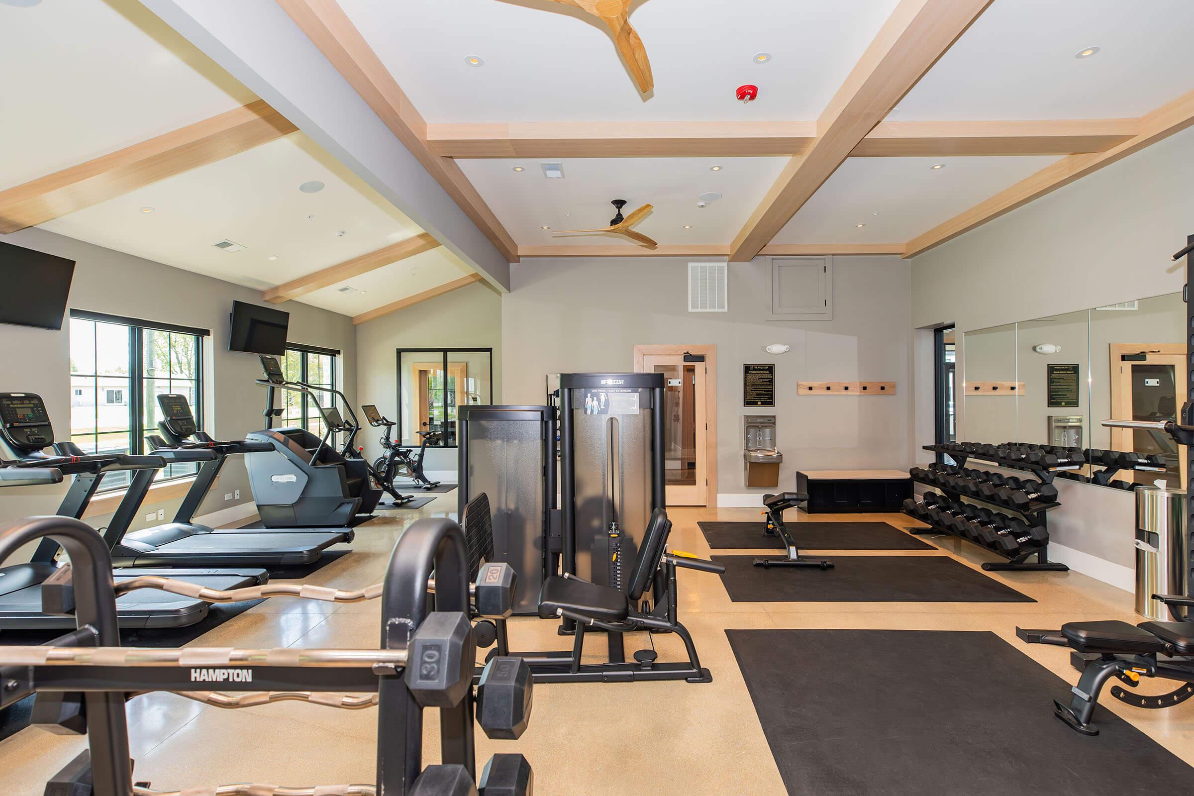 A modern gym interior featuring various exercise equipment, including treadmills, weight machines, and free weights. Large windows allow natural light, and the design incorporates a sleek, contemporary aesthetic with wooden beams and a ceiling fan.