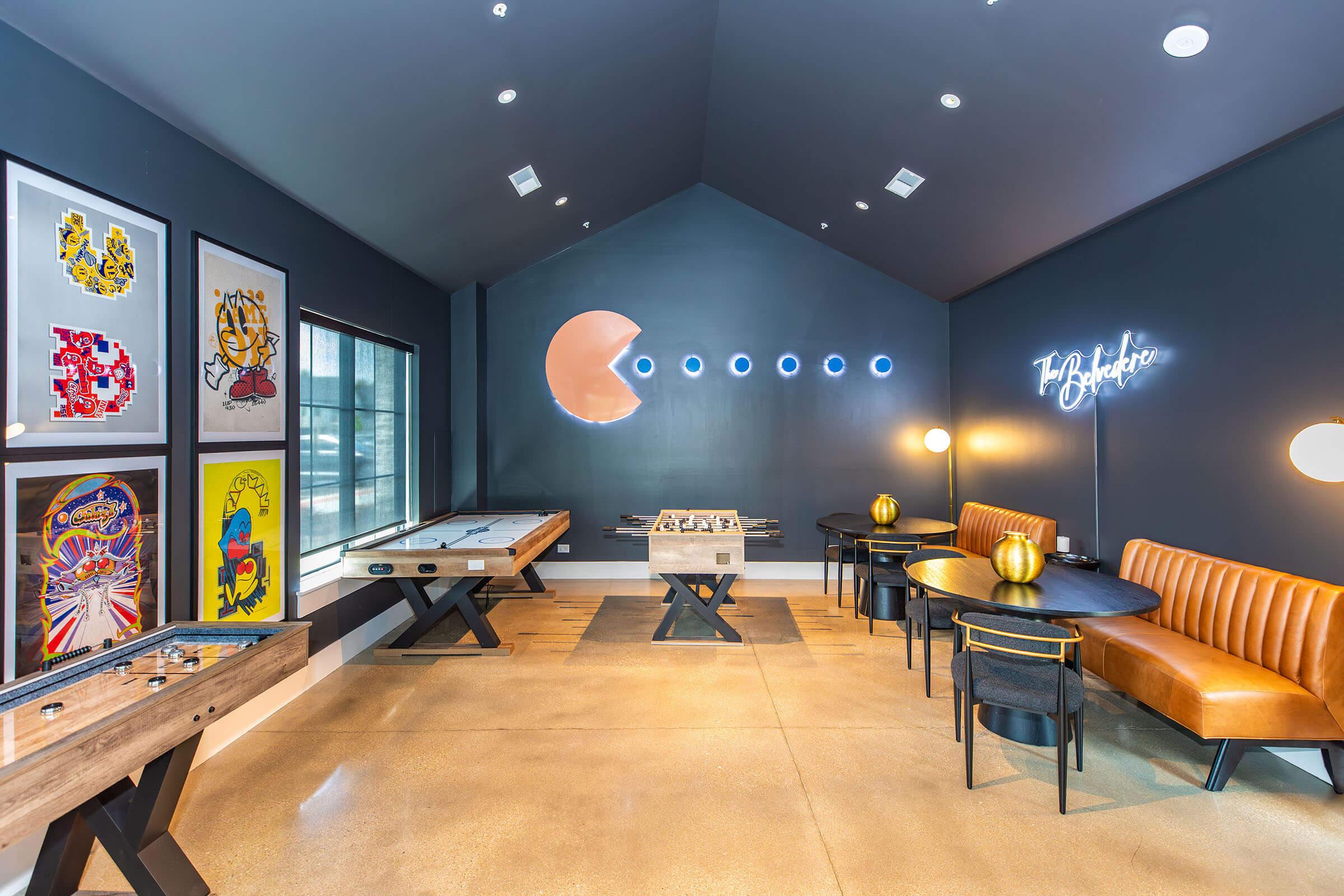 A modern recreational room featuring a foosball table, an air hockey table, and stylish black and orange seating. The walls are adorned with colorful artwork, and a neon sign reads "Be Happy." The space has large windows allowing natural light and a sleek, contemporary design with a high ceiling.