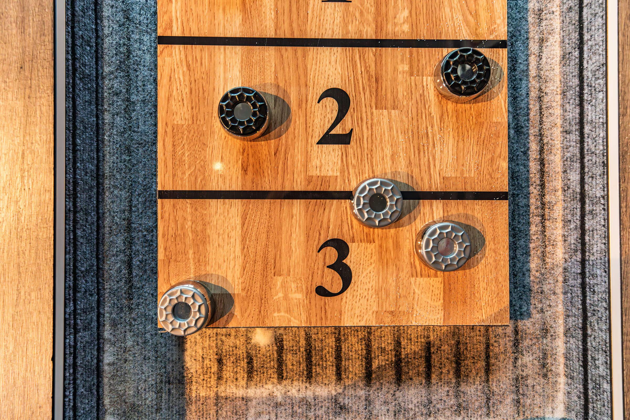 A wooden game board divided into numbered sections with two black and two gray tokens positioned on it. The numbers 2 and 3 are visible, indicating the scoring areas. The board has a textured surface, and the overall setting appears well-lit.
