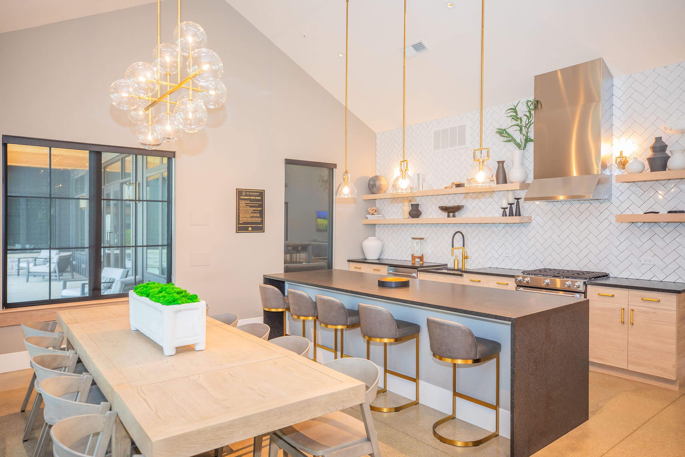 A modern kitchen and dining area featuring a large dining table, sleek bar stools, and a contemporary kitchen layout. The space is bright with natural light, accented by stylish light fixtures, potted plants, and elegant decor elements on shelves, creating a warm and inviting atmosphere.