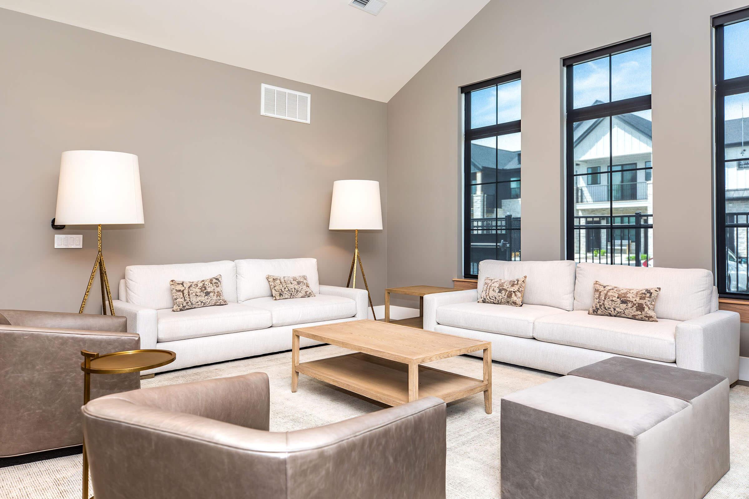 A modern living room featuring two light-colored sofas with decorative pillows, a wooden coffee table, and square armchairs. Bright windows allow natural light, illuminating the space, which has a neutral color palette and minimalist decor. Stylish floor lamps add warmth to the ambiance.