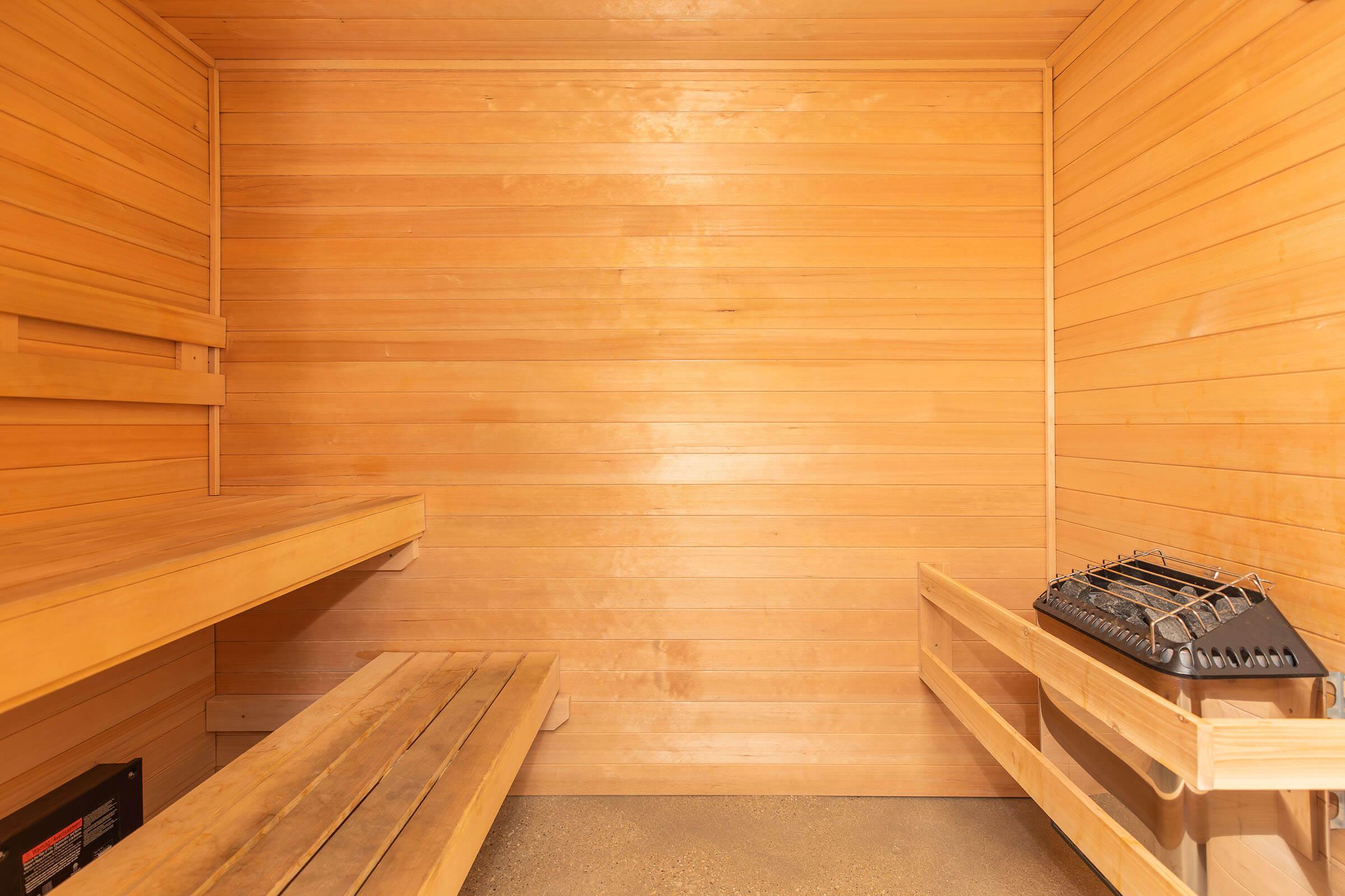 A wooden sauna interior featuring two long benches along the walls and a heater on a shelf. The walls and ceiling are comprised of warm-toned wooden planks, creating a relaxing and cozy atmosphere. The floor is a simple, smooth surface, and the overall lighting is soft and inviting.