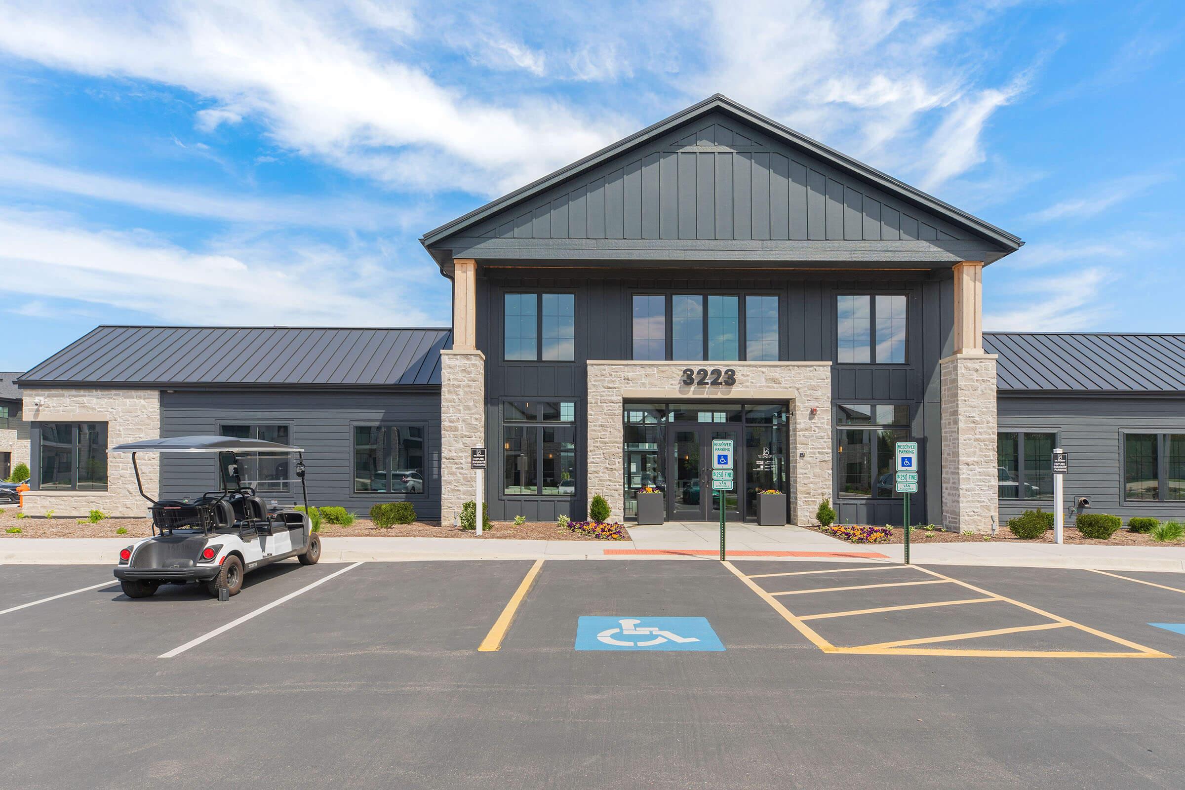 A modern commercial building with a dark gray facade and stone accents, featuring large windows and a welcoming entrance. In the foreground, there is a golf cart parked in a designated area, with accessible parking spaces visible. The sky is clear with a few clouds.