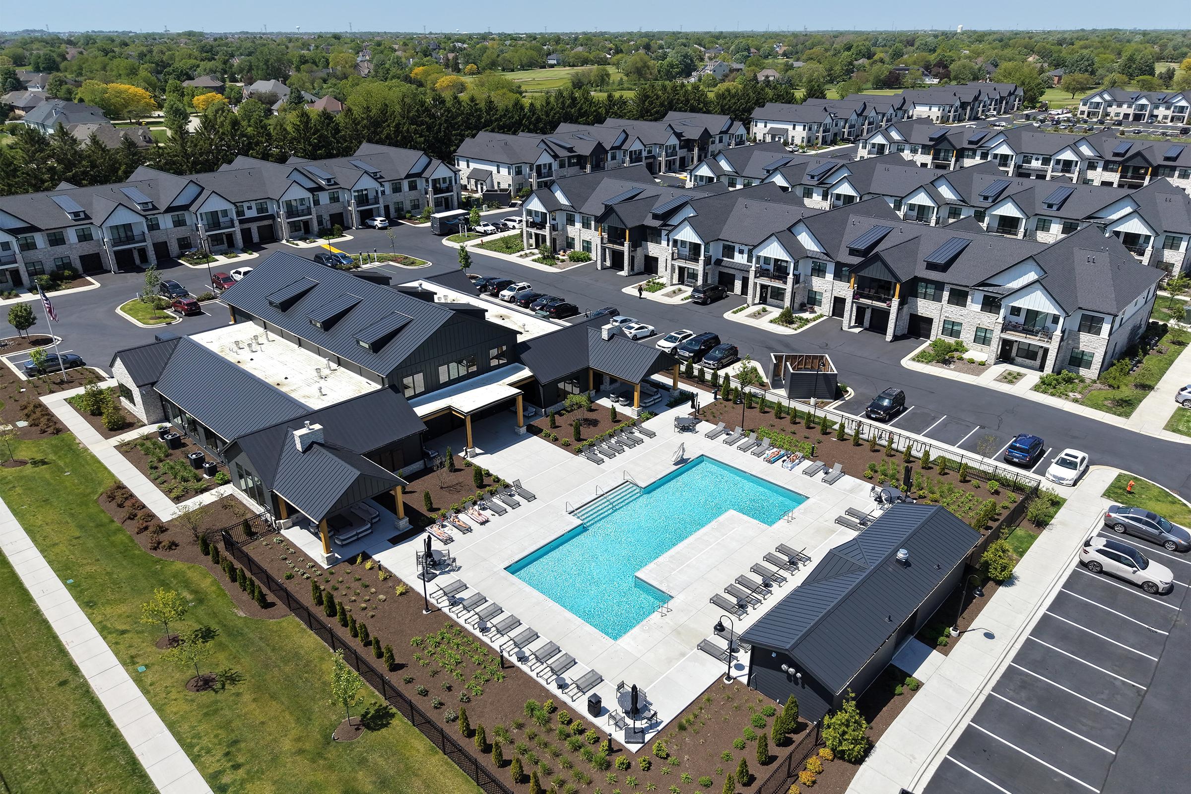Aerial view of a modern residential complex featuring multiple buildings with black roofs. In the foreground, a beautifully landscaped area includes a pool surrounded by lounge chairs, walking paths, and greenery. Parking spaces and several cars are visible, showcasing a suburban atmosphere on a clear day.
