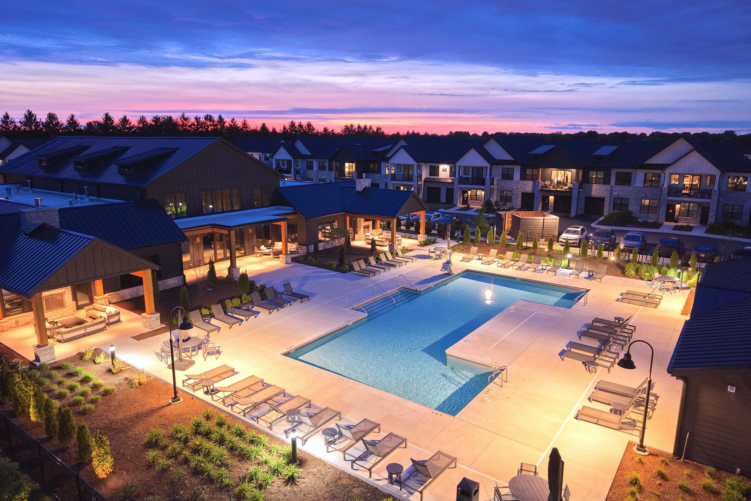 Aerial view of a modern apartment complex showcasing a large outdoor swimming pool surrounded by lounge chairs. The area is illuminated with soft lights, and the sunset creates a colorful sky in the background. Lush landscaping and amenities are visible, adding to the inviting atmosphere.