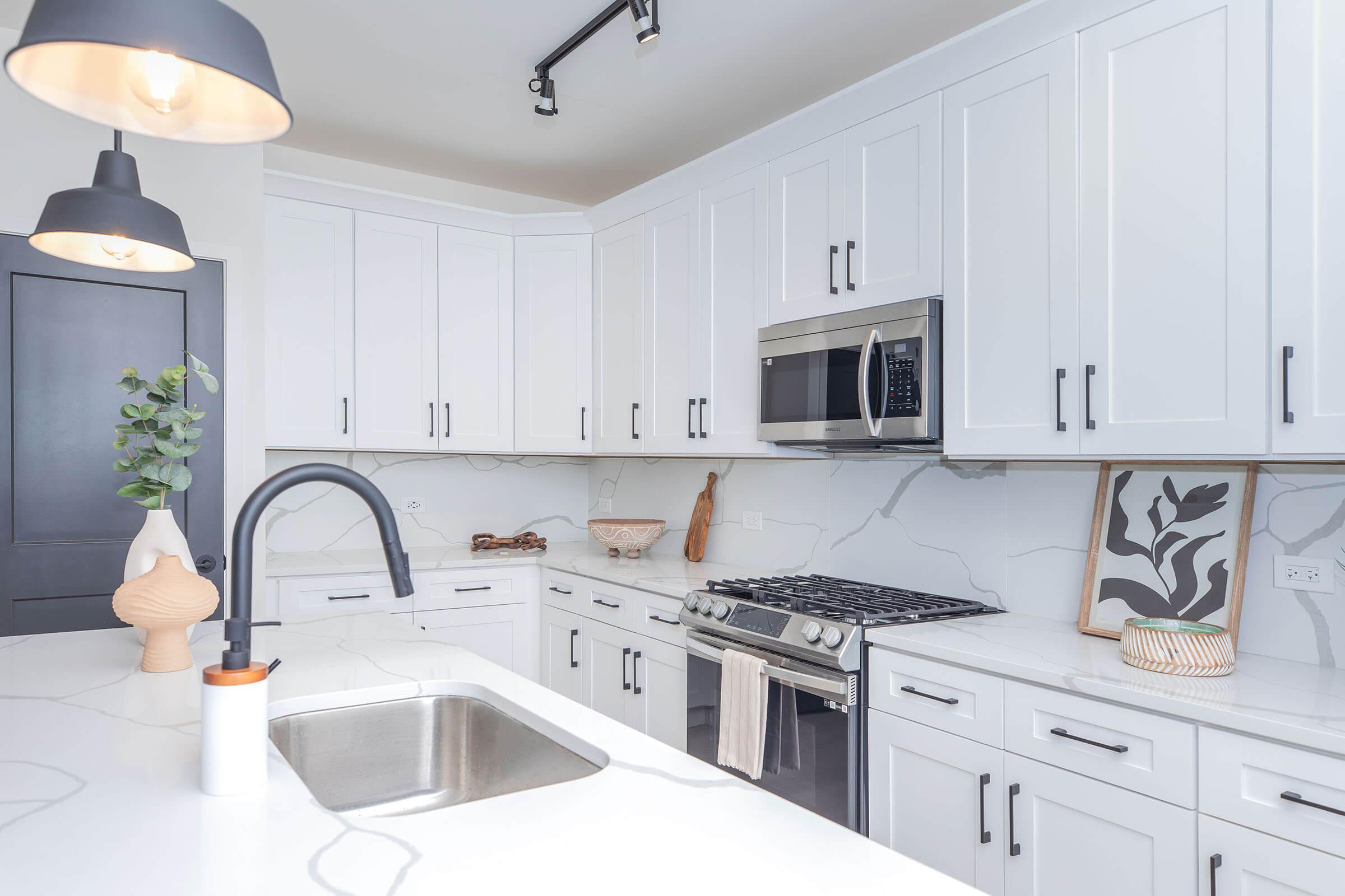A modern kitchen featuring white cabinetry, a stainless steel microwave and gas stove, and a large sink with a sleek faucet. The countertop is made of marble with a marbled design, and there are decorative elements like a vase and artwork on the walls. Pendant lights illuminate the space.