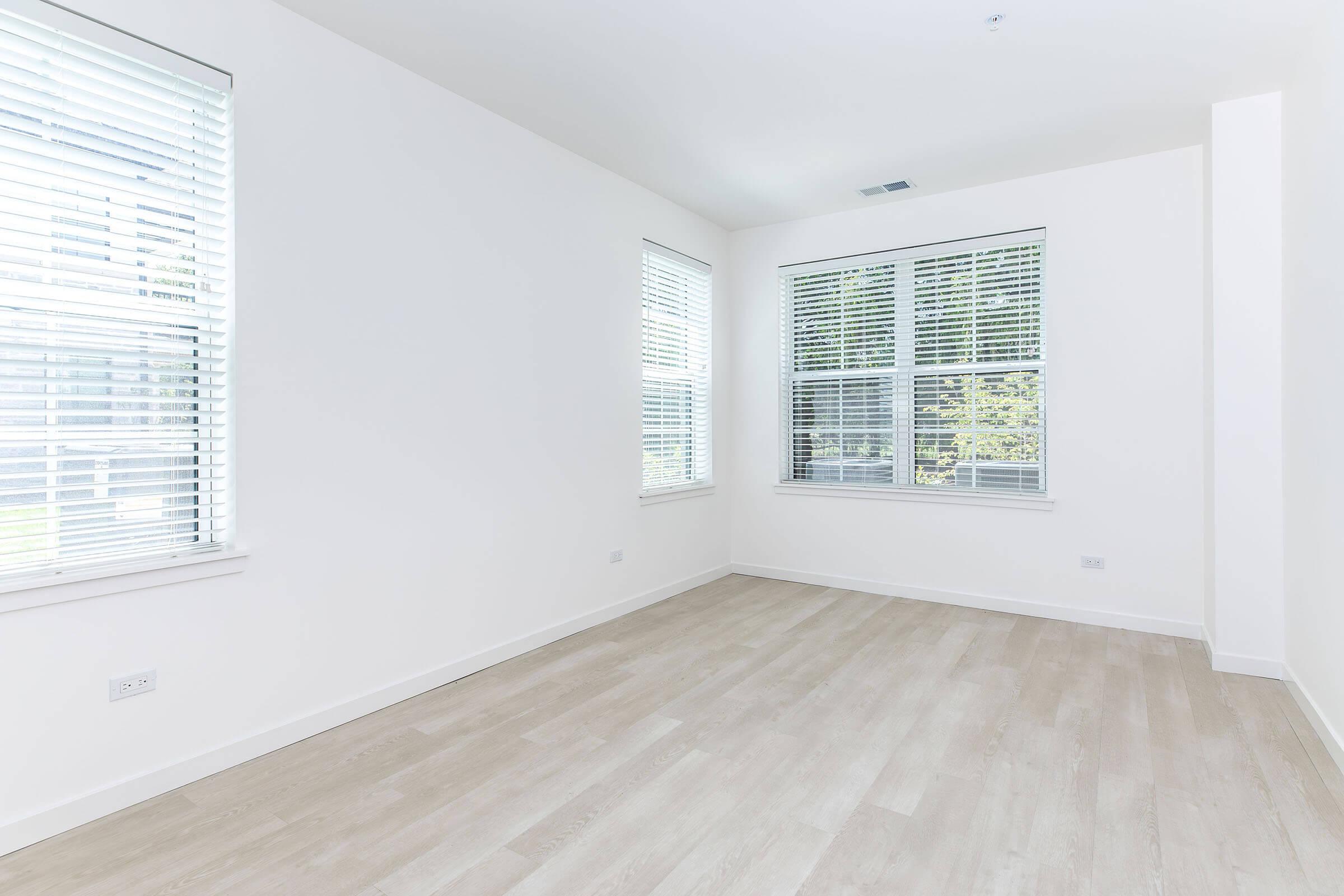 A bright, empty room with white walls and large windows featuring white blinds, allowing natural light to fill the space. The floor is light-colored, possibly wood or laminate, and there are no furnishings, emphasizing the room's openness and simplicity.