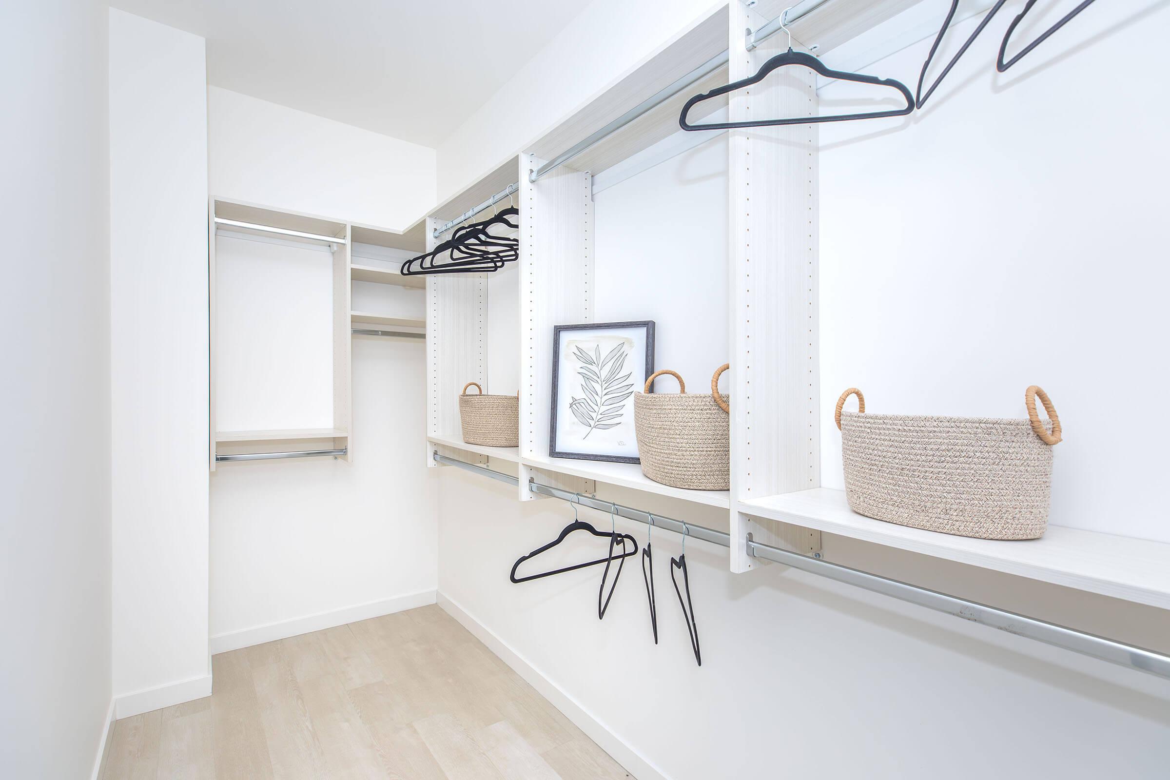 A spacious, minimalist closet featuring white shelving and hangers. On the shelves, there are three woven storage baskets and a framed leaf print. The walls are clean and bright, with a light wooden floor, creating an organized and appealing storage space.