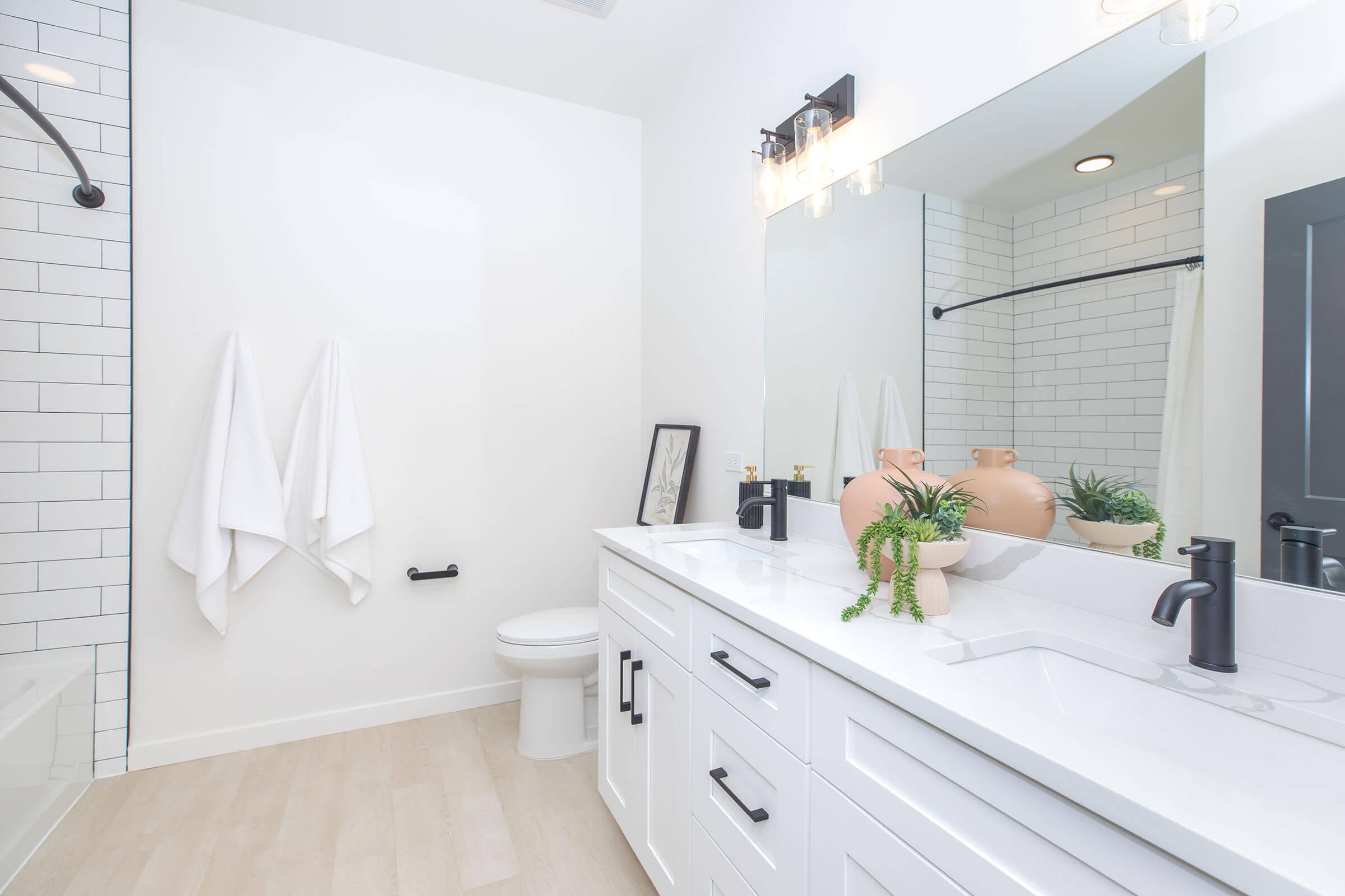 Modern bathroom featuring a white vanity with double sinks, stylish fixtures, and a large mirror. A shower area with black accents is visible, along with two white towels hanging on the wall. Decorative plants and ceramics add a touch of elegance to the minimalist design. Neutral color palette enhances the spacious feel.