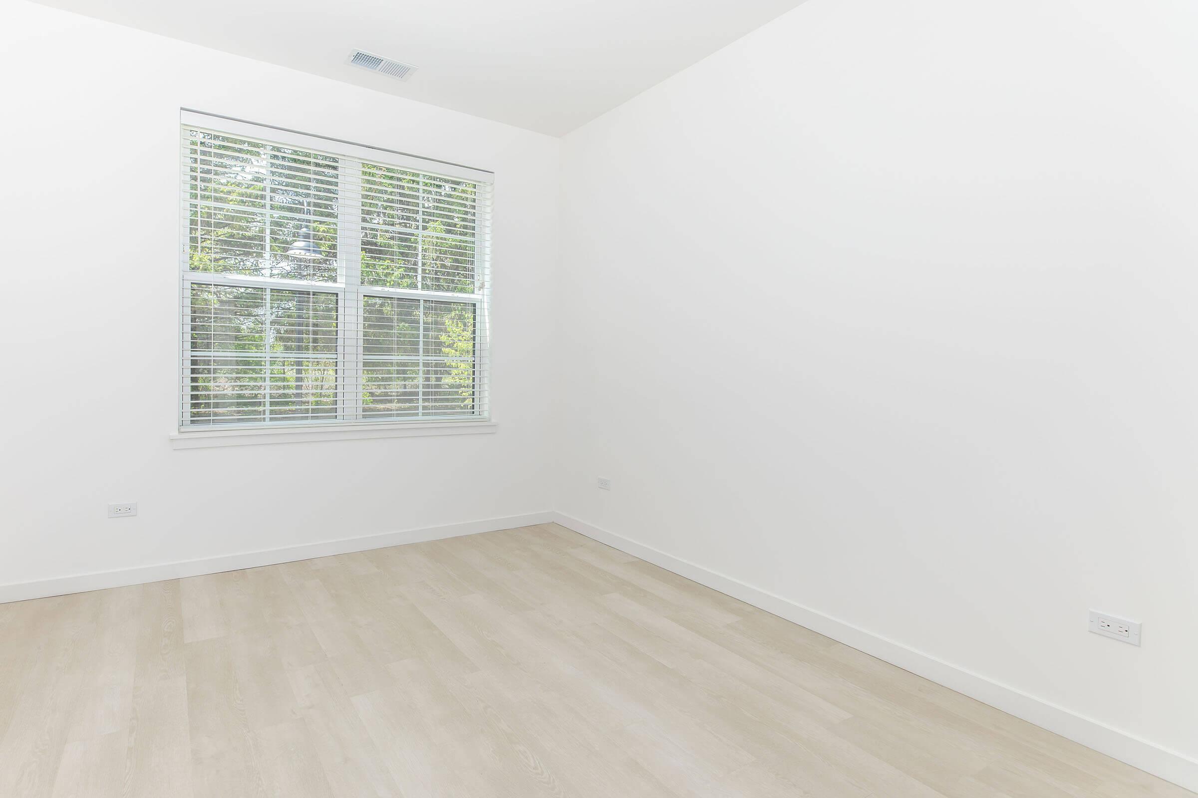 A bright, empty room with light-colored walls and wooden flooring. There is a large window with blinds that lets in natural light, illuminating the space. The room has a simple, clean aesthetic, suitable for various purposes such as an office or relaxation area.