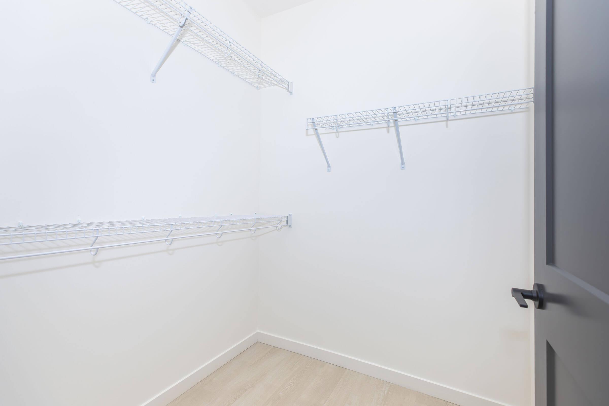 Empty closet with white walls and light wooden flooring. There are several wire shelving units mounted on the walls, providing storage space. The closet has a dark door with a handle, and overall, it appears neat and organized, ready for use.