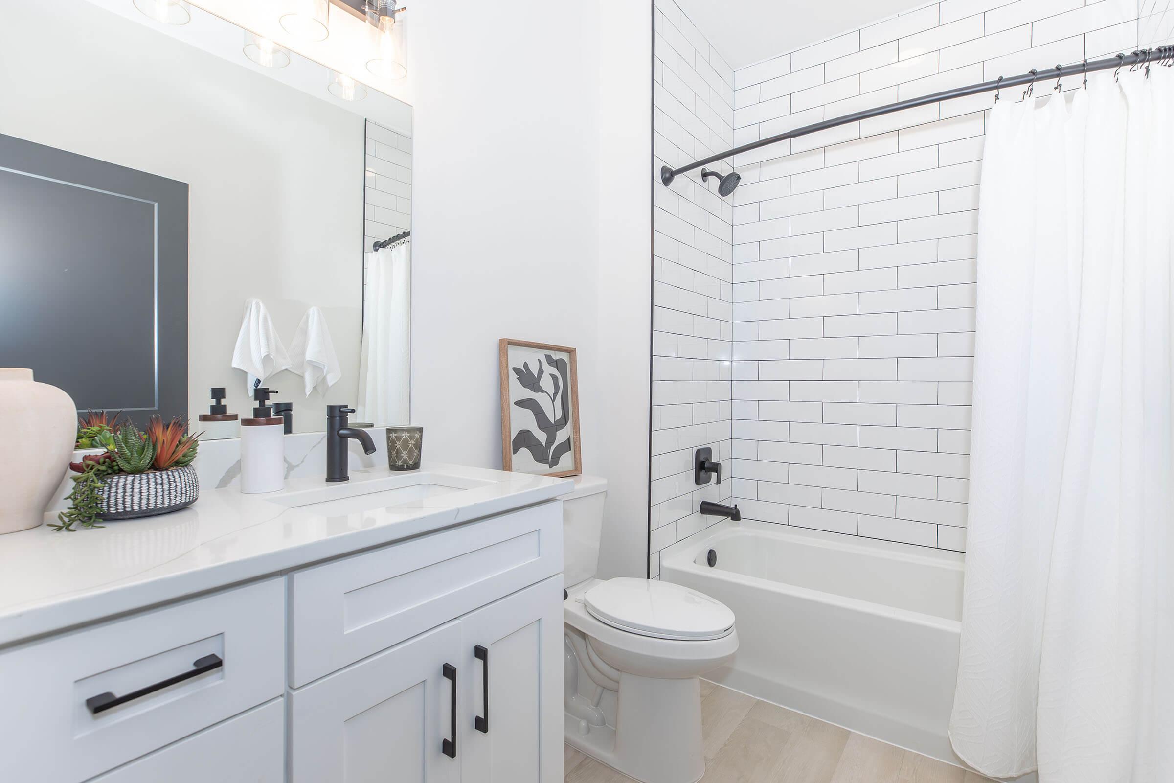 Modern bathroom featuring a white tiled wall, a bathtub with a shower curtain, a sleek vanity with a sink, and stylish decor items like a framed print and potted plants. Bright lighting illuminates the space, creating a clean and inviting atmosphere.
